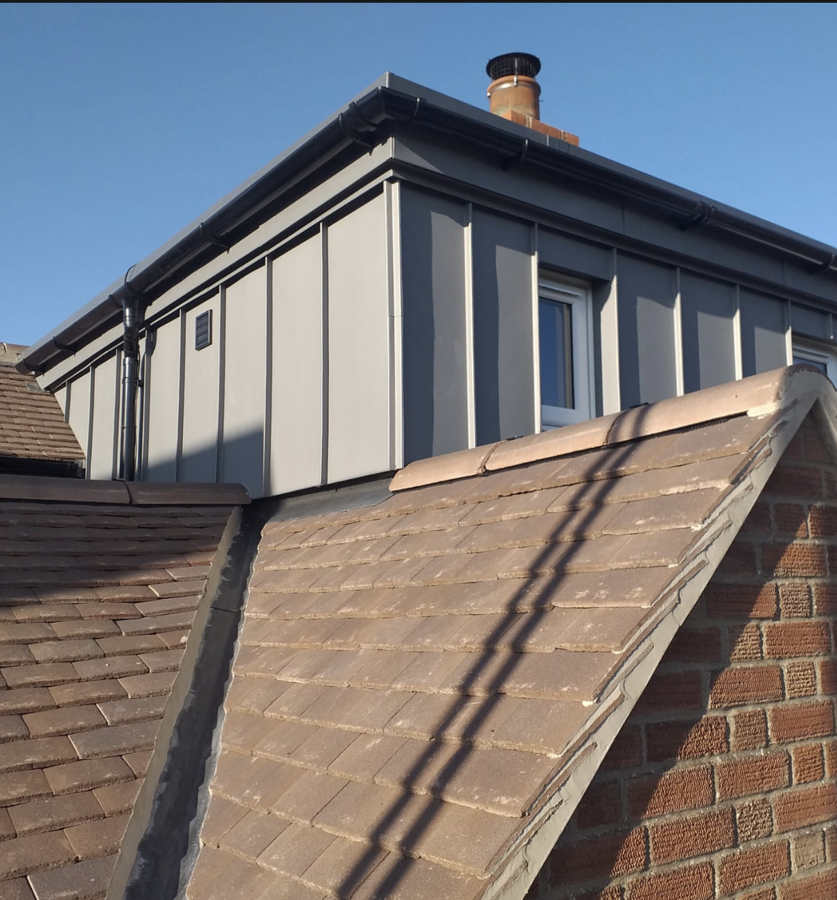 Close-up of a residential roof with brown tiles and a small dormer window with grey cladding under a clear blue sky.
