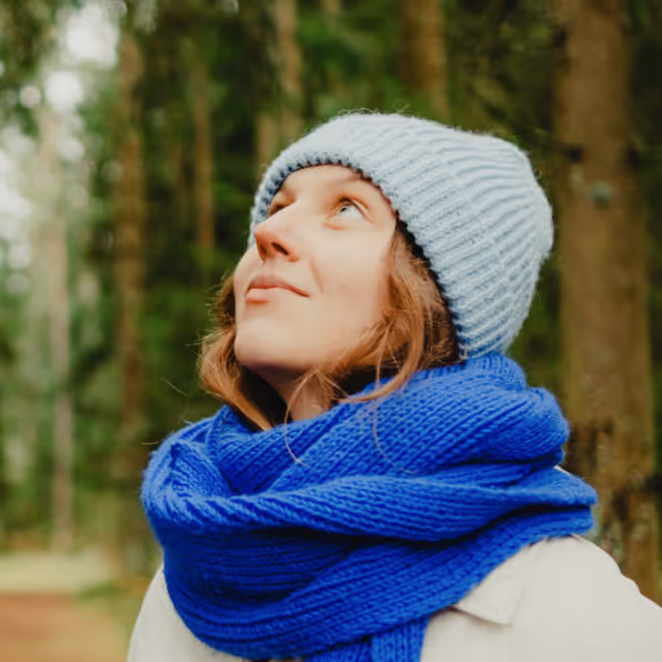 Young woman wearing a white knit hat and a thick blue scarf, looking up while standing in a forest.