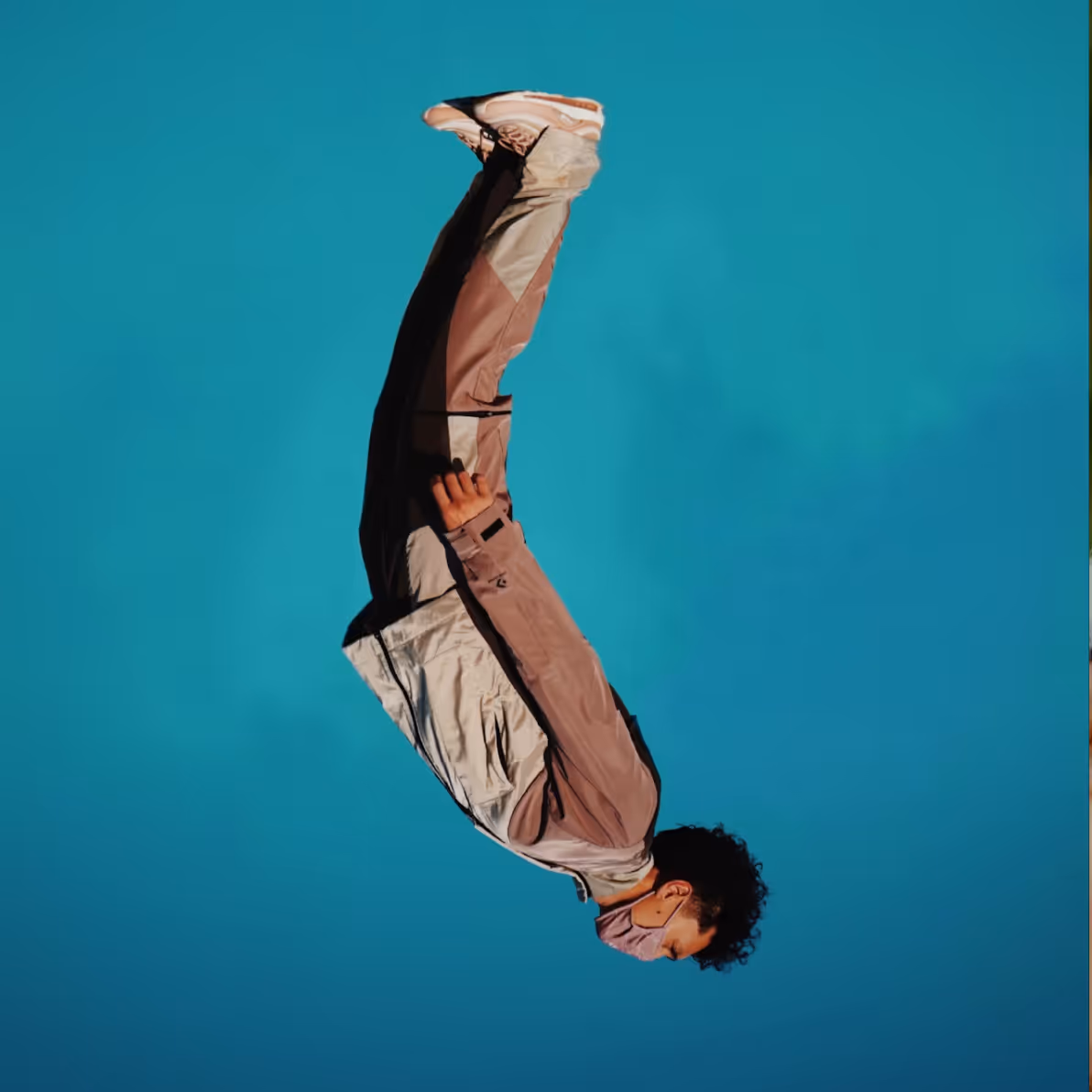 Person in light-colored jacket, pants, sneakers, and face mask, captured mid-backflip against a clear blue sky.