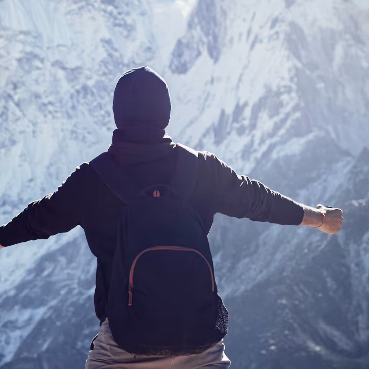 Person wearing a black hoodie and backpack with arms outstretched facing snow-covered mountain peaks.