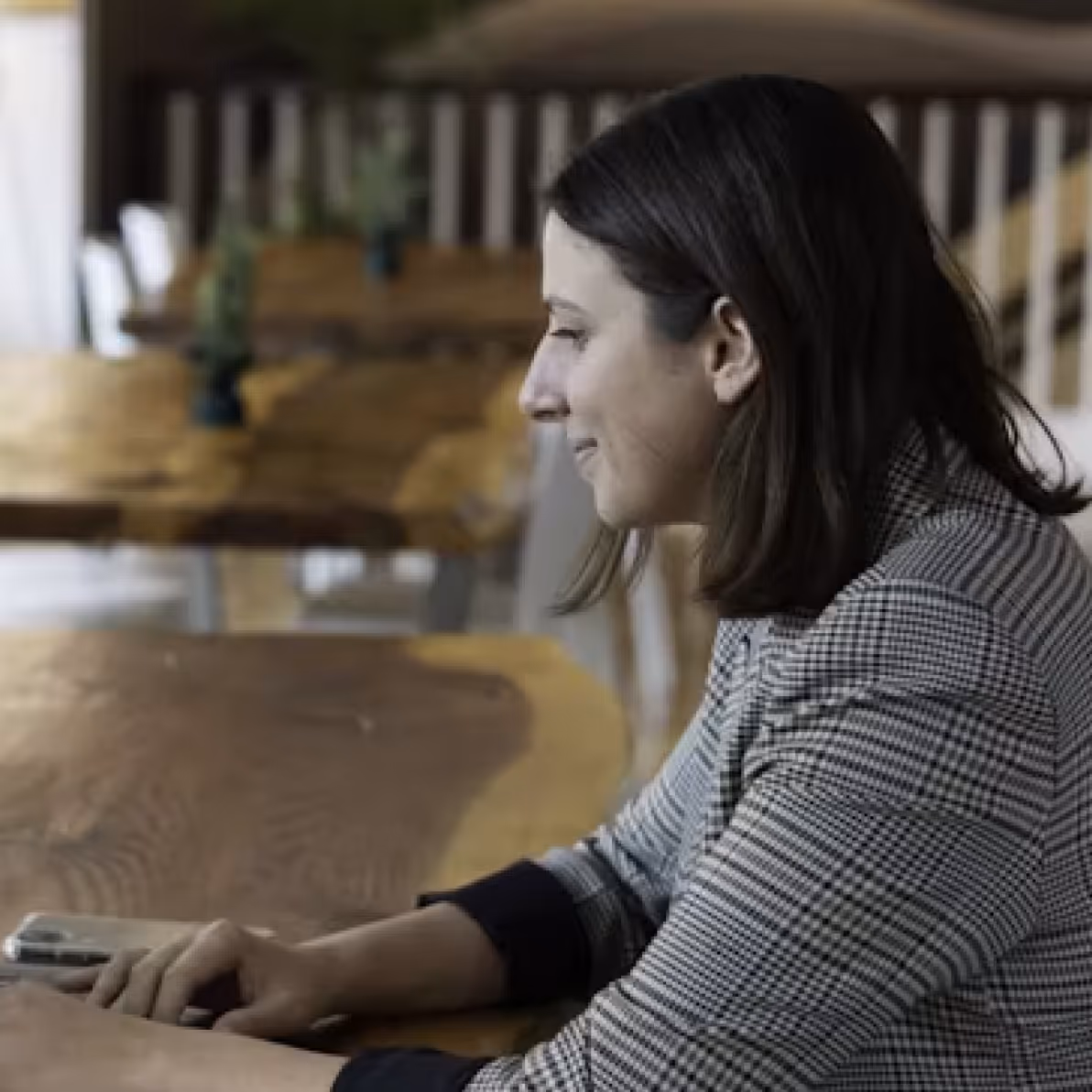 Woman with shoulder-length dark hair in a checkered blazer sitting at a wooden table, looking at a smartphone.