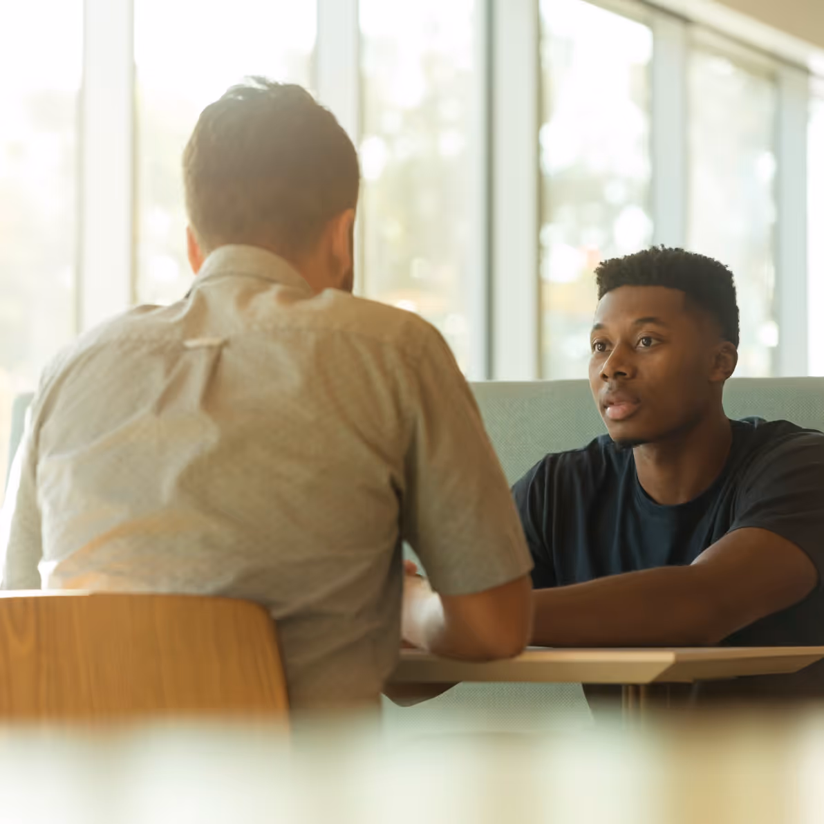 Two men engaged in a serious conversation while seated at a table in a bright room with large windows.