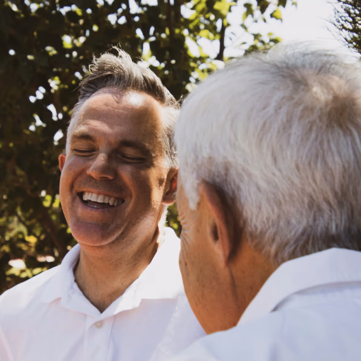 Two men outdoors, one younger man smiling with eyes closed and an older man with gray hair seen from behind.