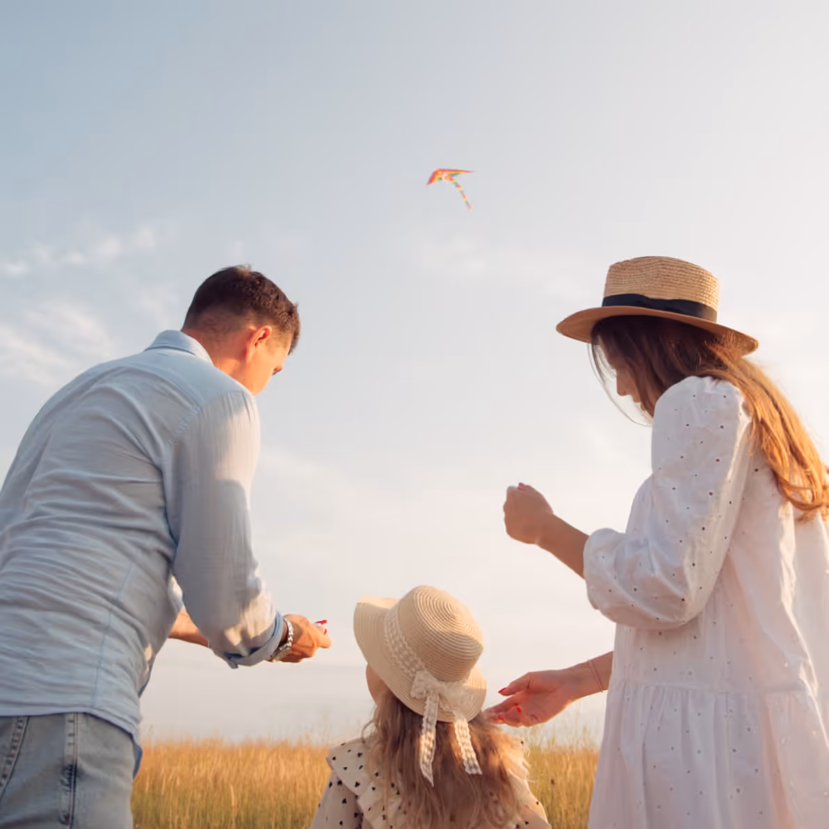 Young family of two adults and a child flying a colorful kite in a field at sunset.