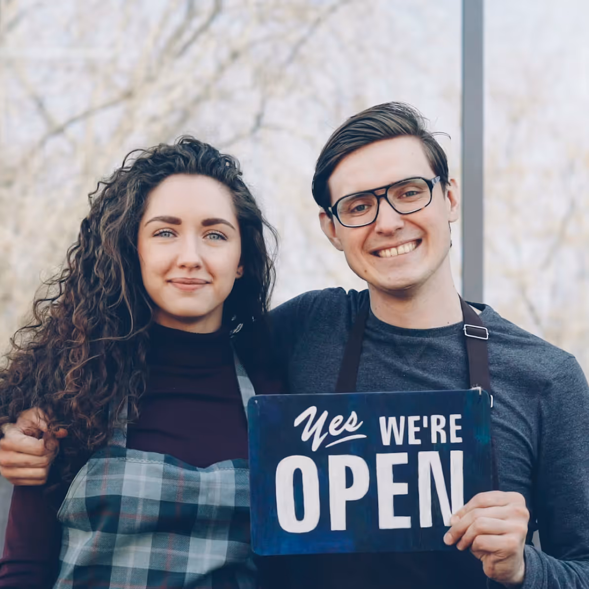 Smiling man and woman wearing aprons holding a sign that says 'Yes we're open' outside.