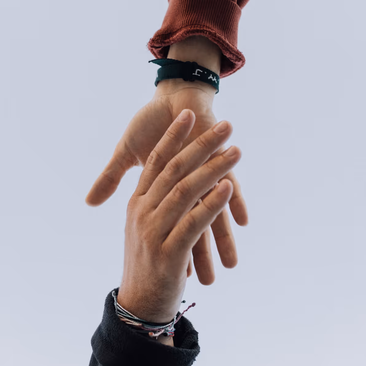 Two hands holding each other against a plain light background, one wearing a green wristband and the other multiple string bracelets.