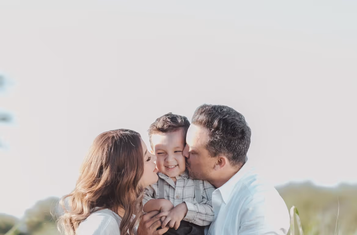 Smiling young boy being kissed on the cheeks by a woman and a man outdoors.