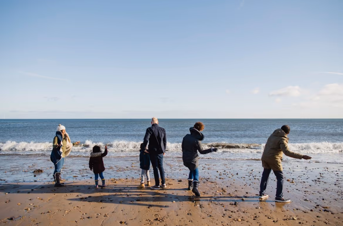 Six people on a beach dressed in winter clothes, facing the sea with waves under a clear sky.