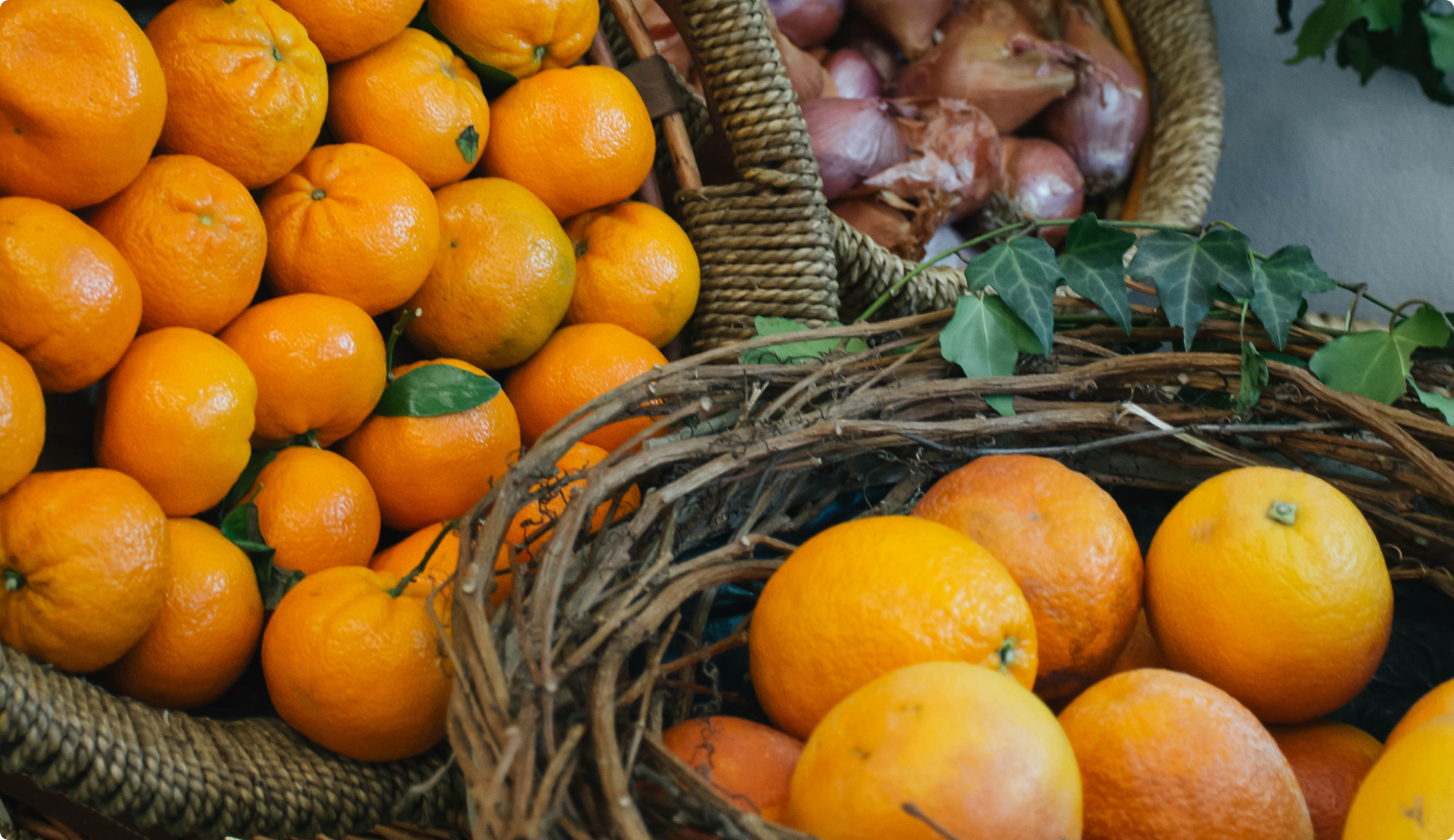 Baskets filled with bright orange mandarins and a separate basket of shallots, with green leaves and twigs nearby.
