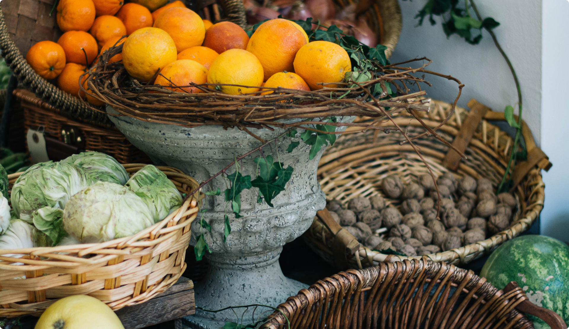 Baskets of fresh produce including oranges, cabbages, walnuts, and a watermelon displayed around a stone pedestal with a rustic vine wreath holding oranges.