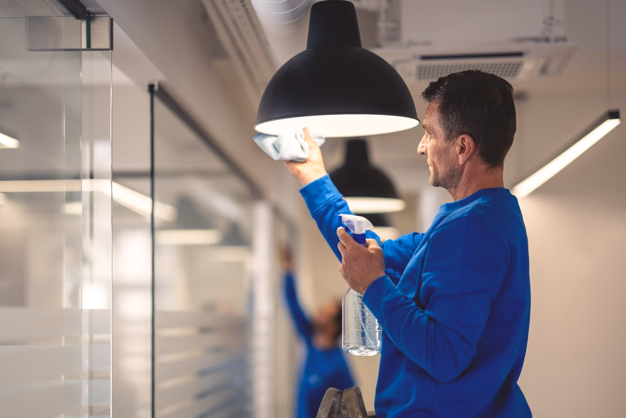 Man in blue cleaning a black ceiling lamp with a cloth and spray bottle in a modern office setting.