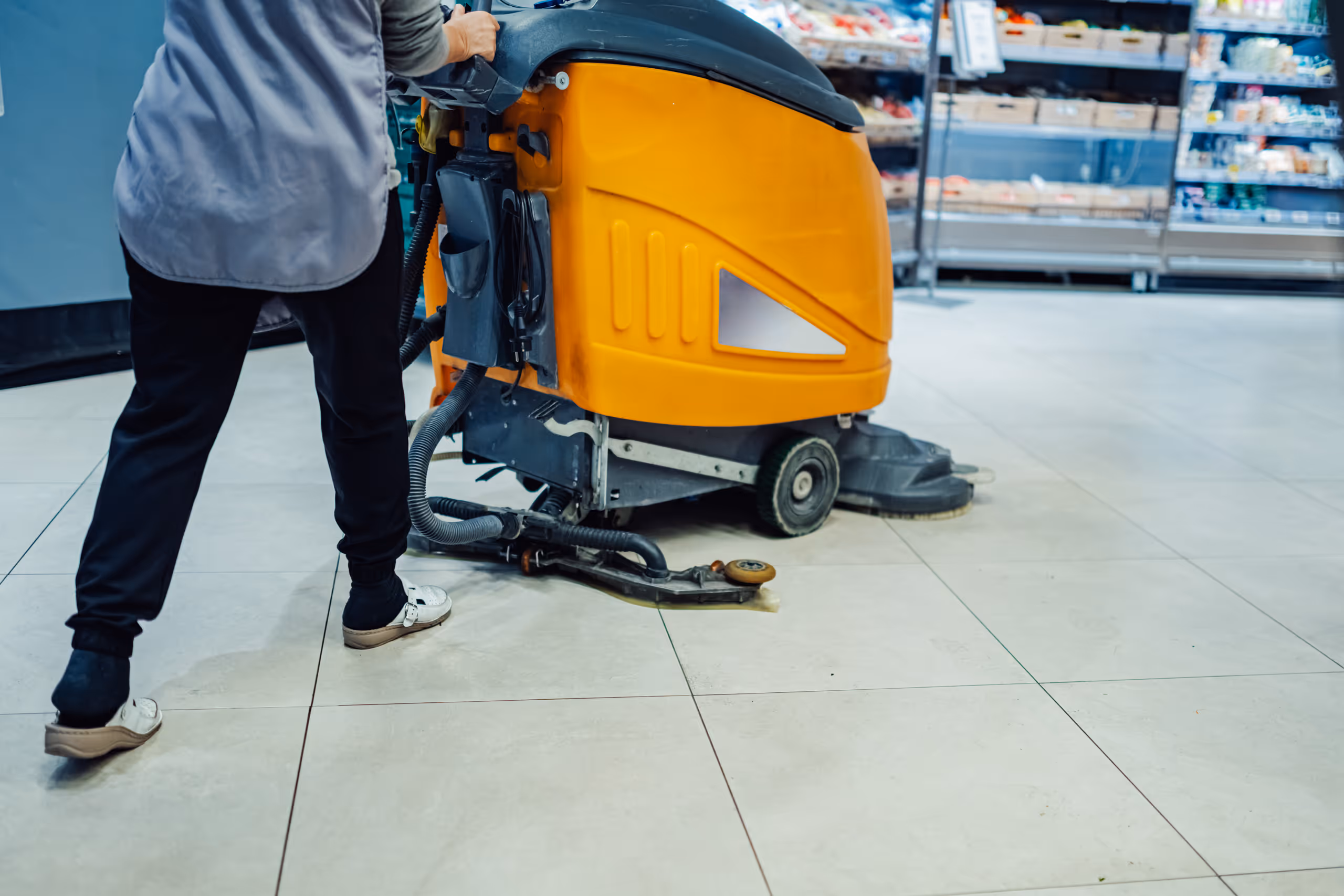 Person operating an orange floor cleaning machine on tiled floor in a store.