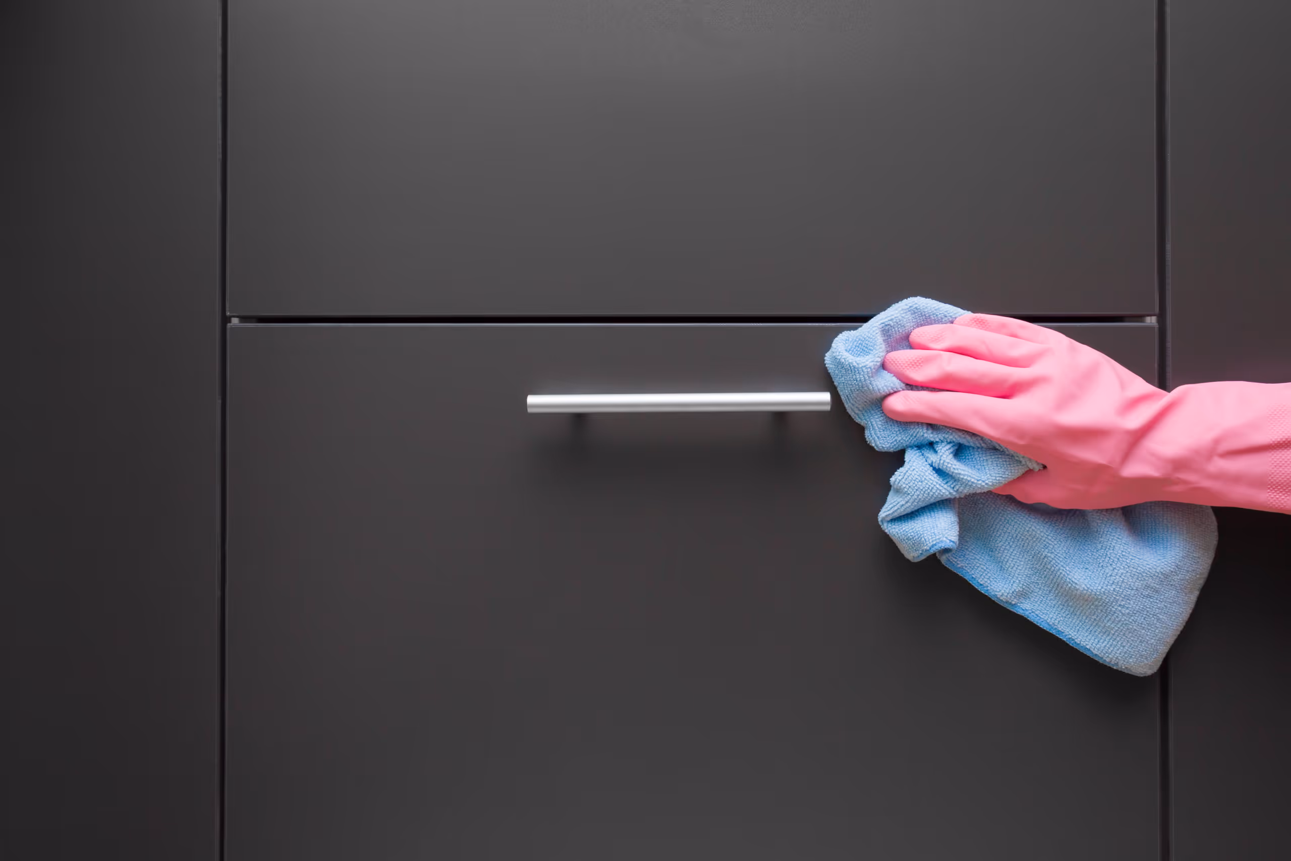Hand wearing pink glove cleaning a dark cabinet handle with a blue cloth.