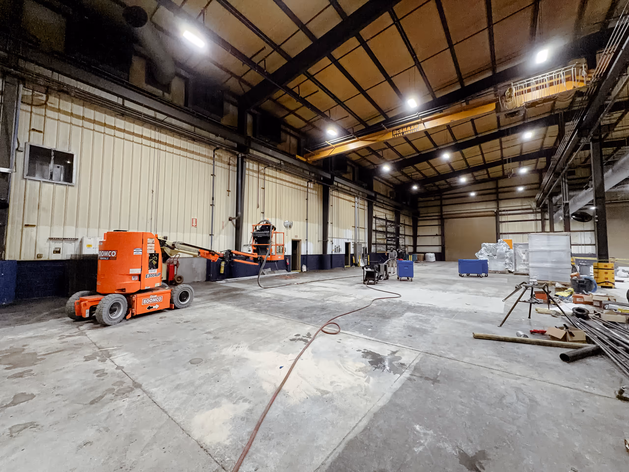Large industrial warehouse interior with orange boom lift and scattered equipment on concrete floor.