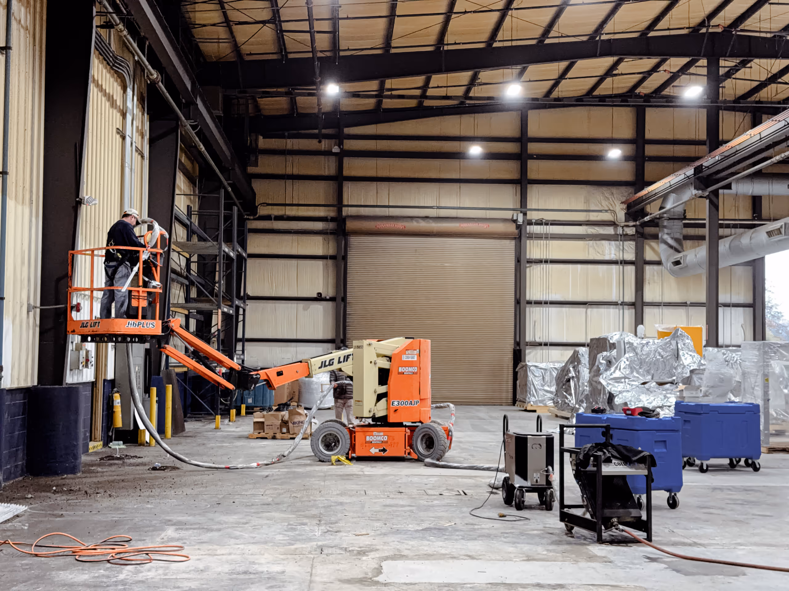 Worker on a JLG lift inside a large industrial warehouse performing maintenance or installation work near the ceiling.