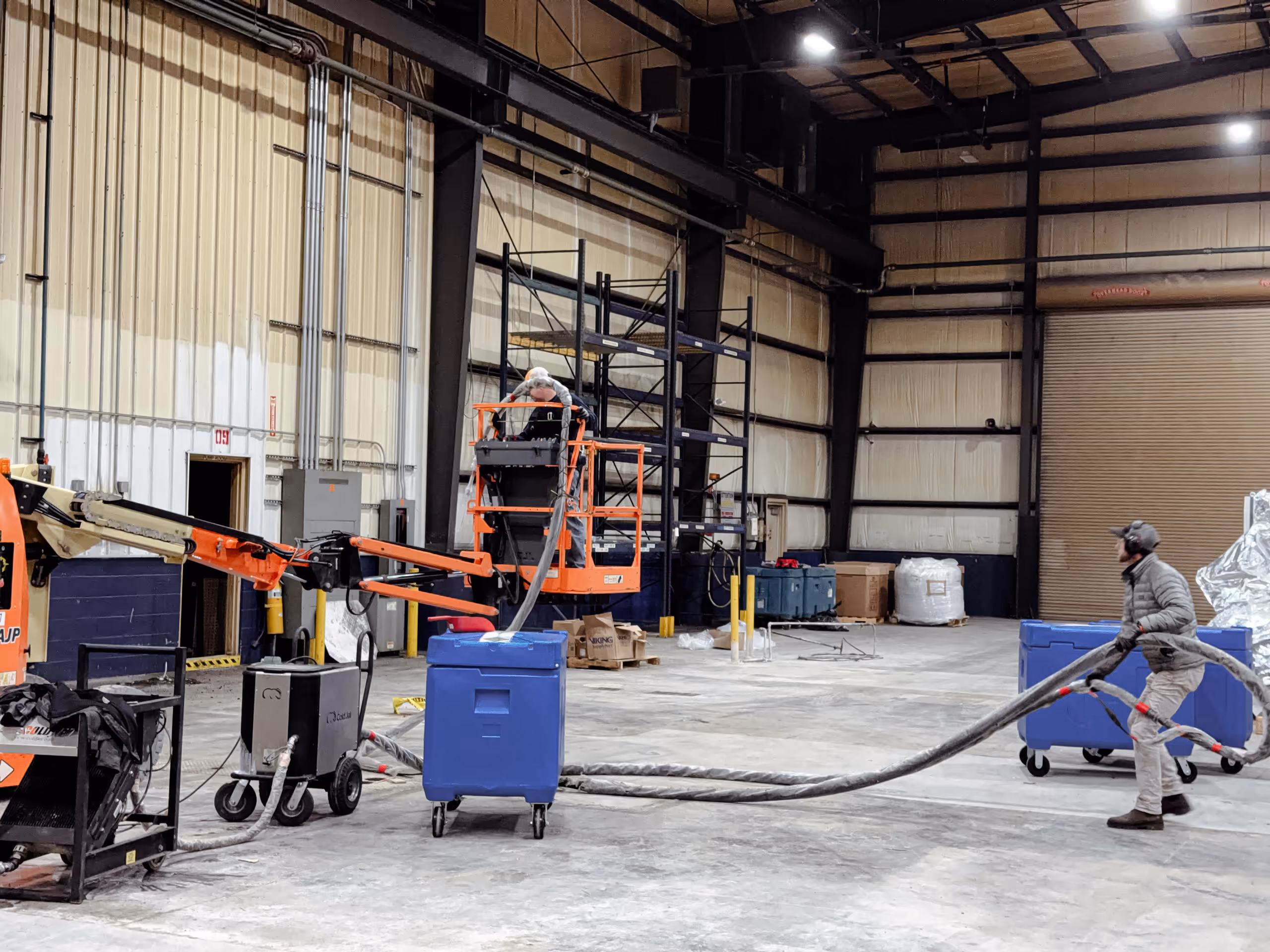 Two workers in protective gear using ice blasting equipment inside a large industrial warehouse.