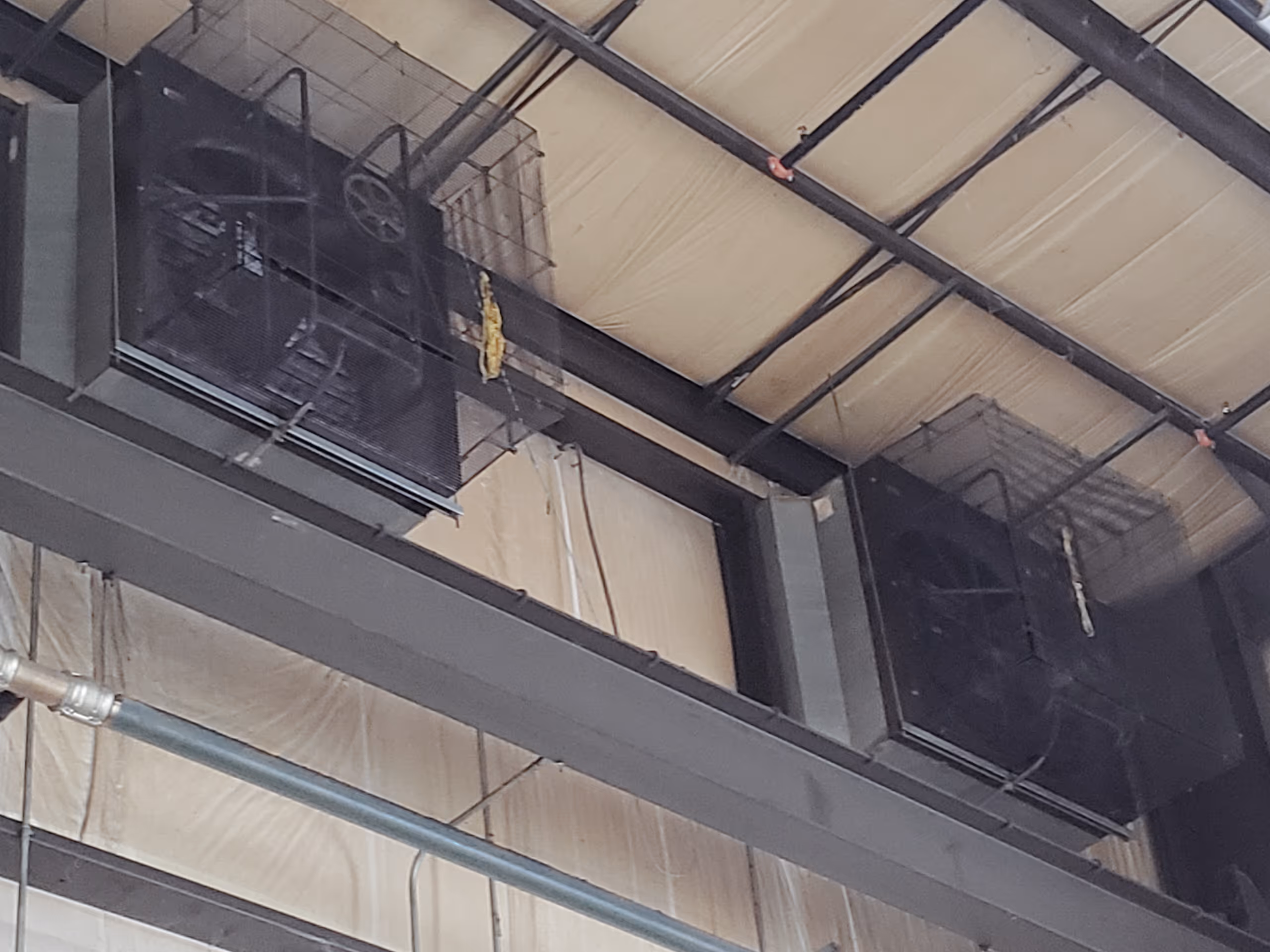 Two large industrial ceiling fans with protective cages mounted on a metal beam inside a warehouse or factory with insulated walls and ceiling.