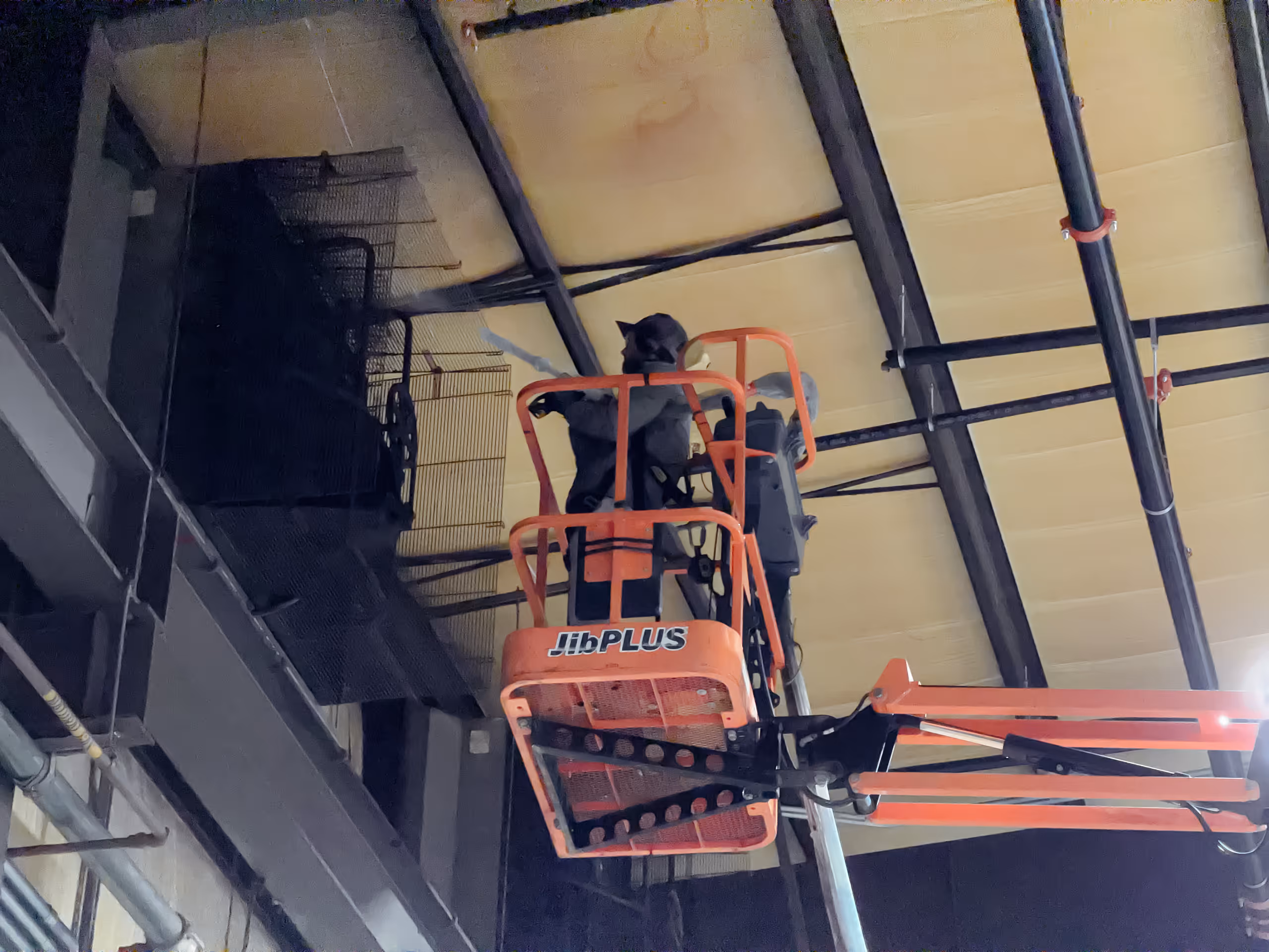 Worker in black attire operating an orange JibPLUS aerial lift inside an industrial building near the ceiling.