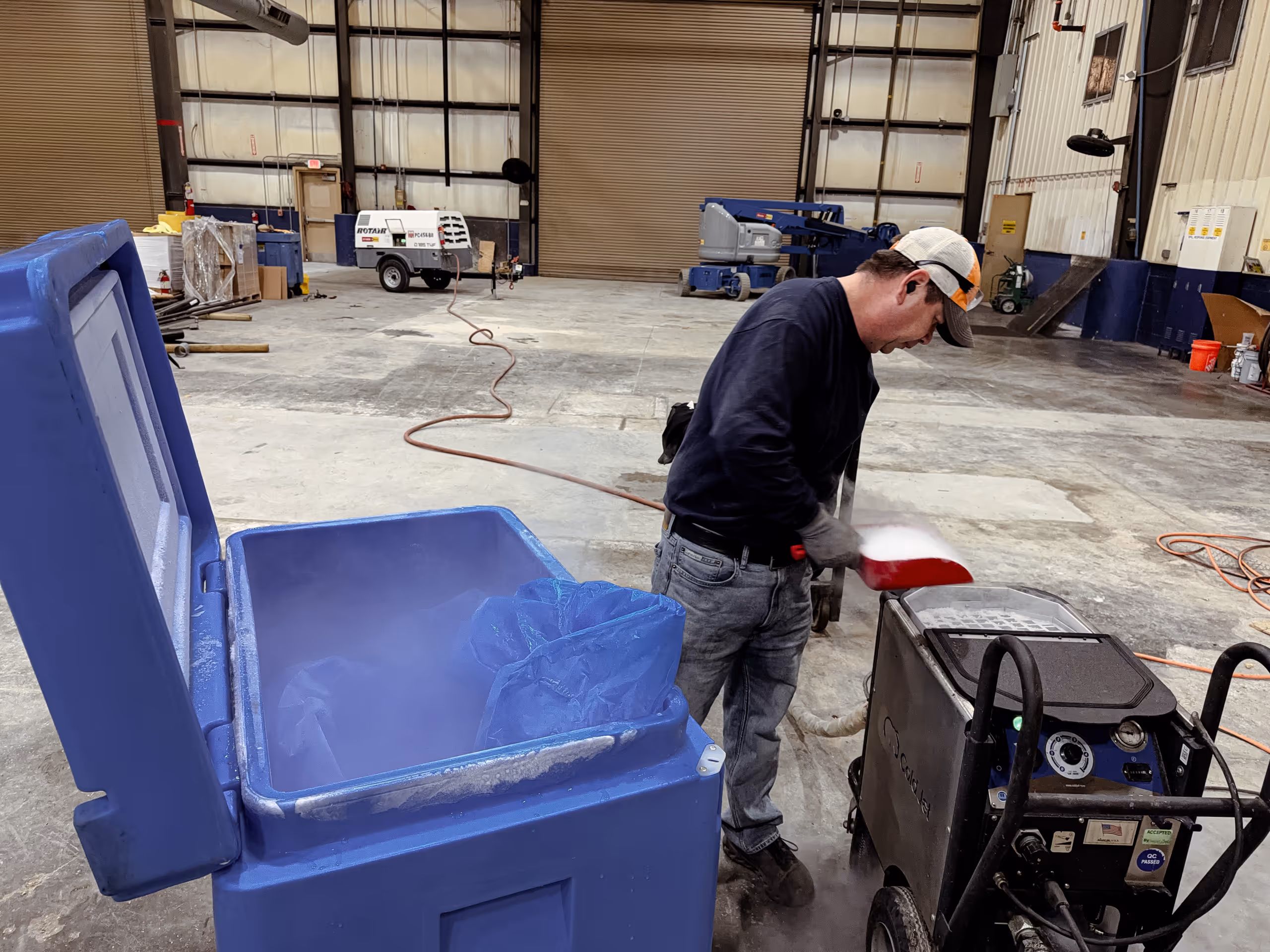 Man wearing a cap and gloves scooping dry ice from a blue container in a large industrial warehouse.