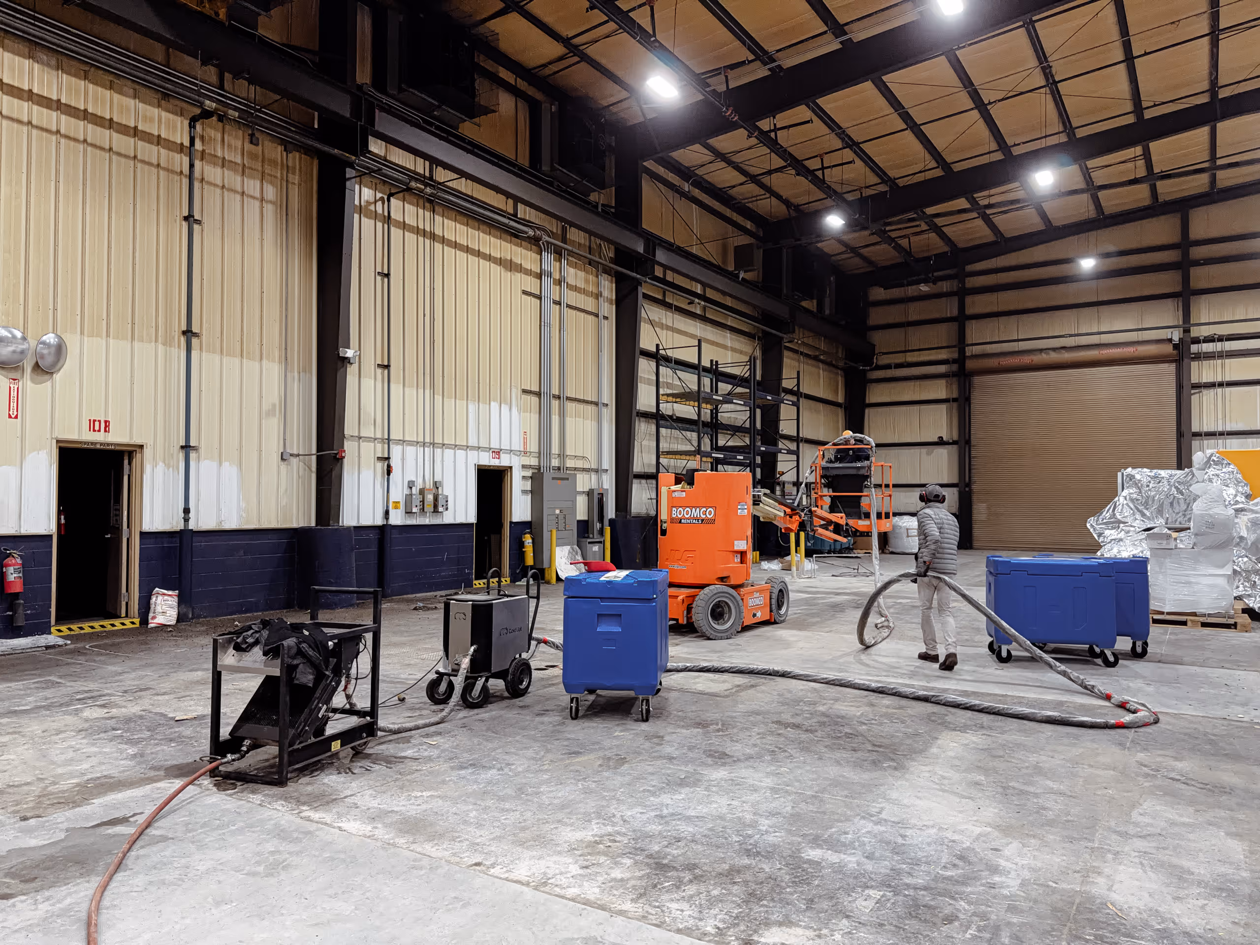 Person operating equipment inside a large industrial warehouse with high ceilings and an orange boom lift.