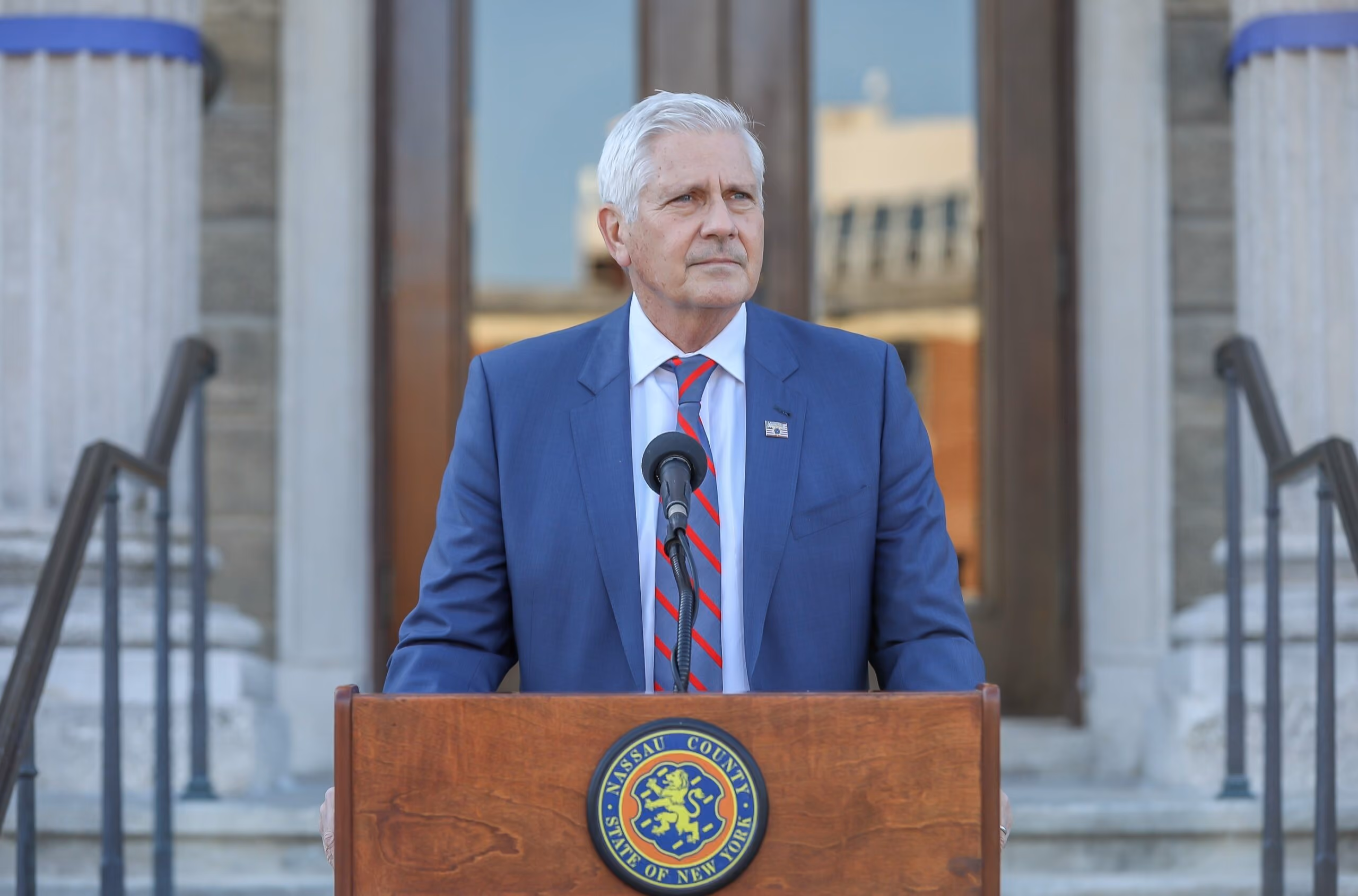 Man in blue suit and striped tie standing at a podium with Nassau County seal and microphone.