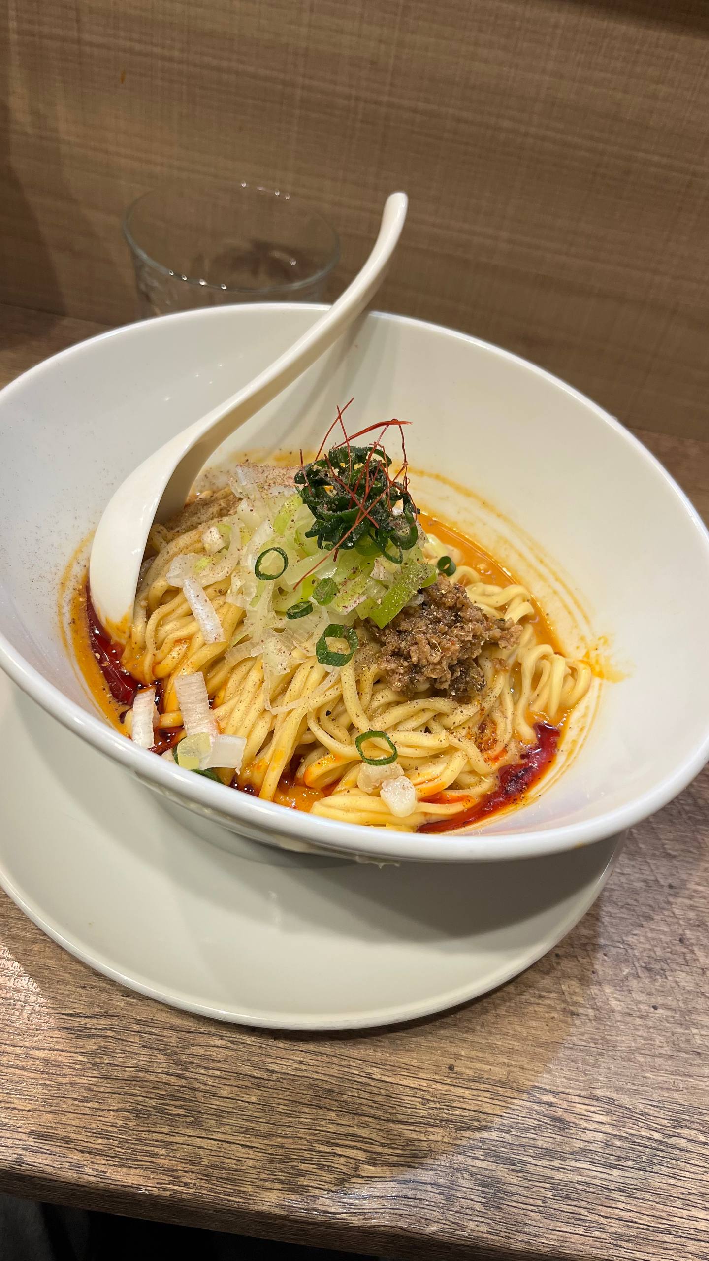 Bowl of spicy ramen noodles topped with minced meat, chopped green onions, sliced white onions, seaweed, and chili oil with a white spoon resting inside.