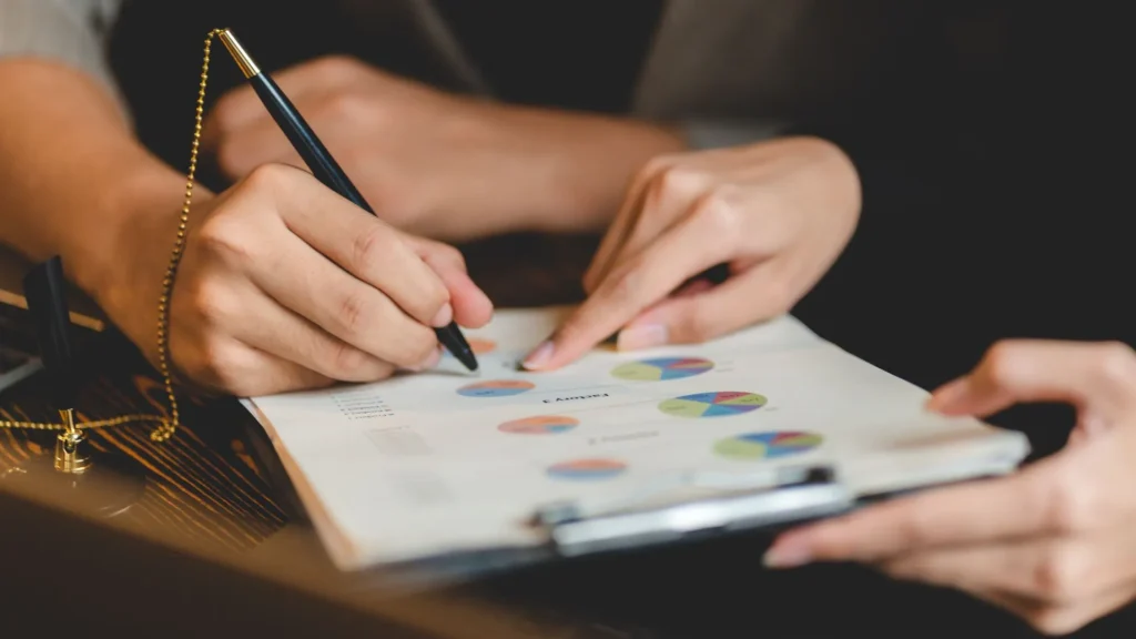 Two people are discussing some charts on a clipboard. One of them is holding a pen.
