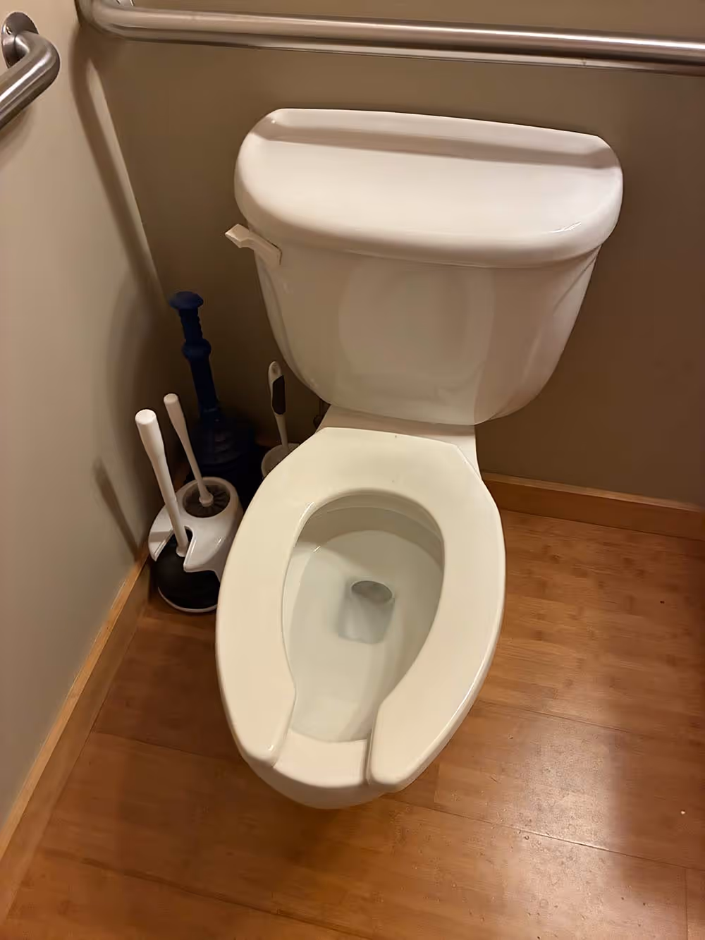 White toilet with open lid in a bathroom corner with wooden floor and multiple toilet plungers beside it.