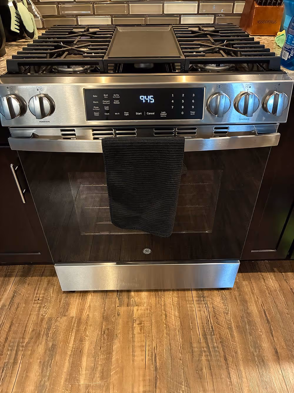 Stainless steel gas stove with black grates and digital clock displaying 9:45 with a black oven mitt hanging on the oven door handle.