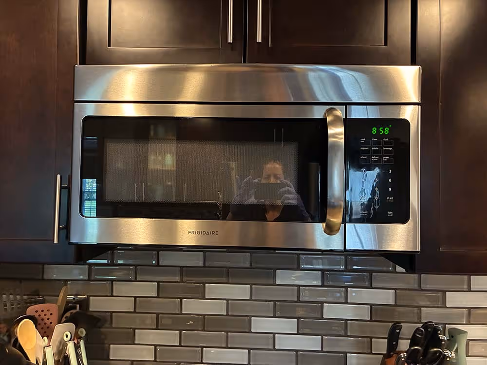 Stainless steel Frigidaire microwave oven mounted above a kitchen stove with dark wood cabinets and grey subway tile backsplash.