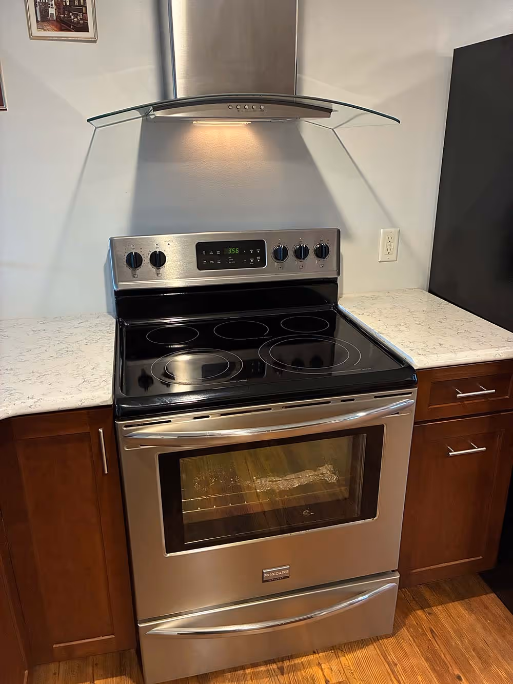 Stainless steel electric stove with glass cooktop and vent hood in a kitchen with wooden cabinets and light-colored countertops.