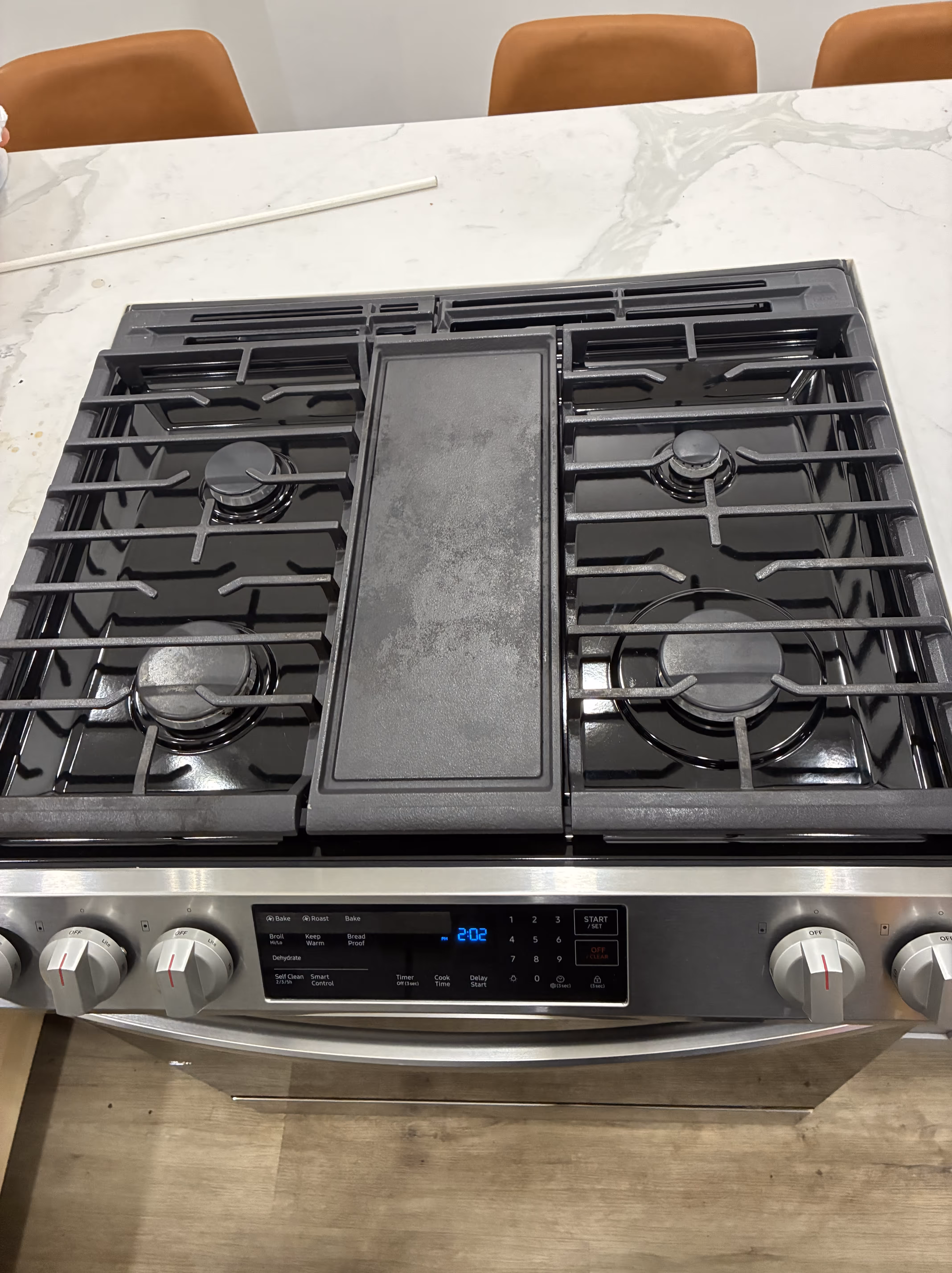 Stainless steel gas stove with black grates and digital clock displaying 9:45 with a black oven mitt hanging on the oven door handle.