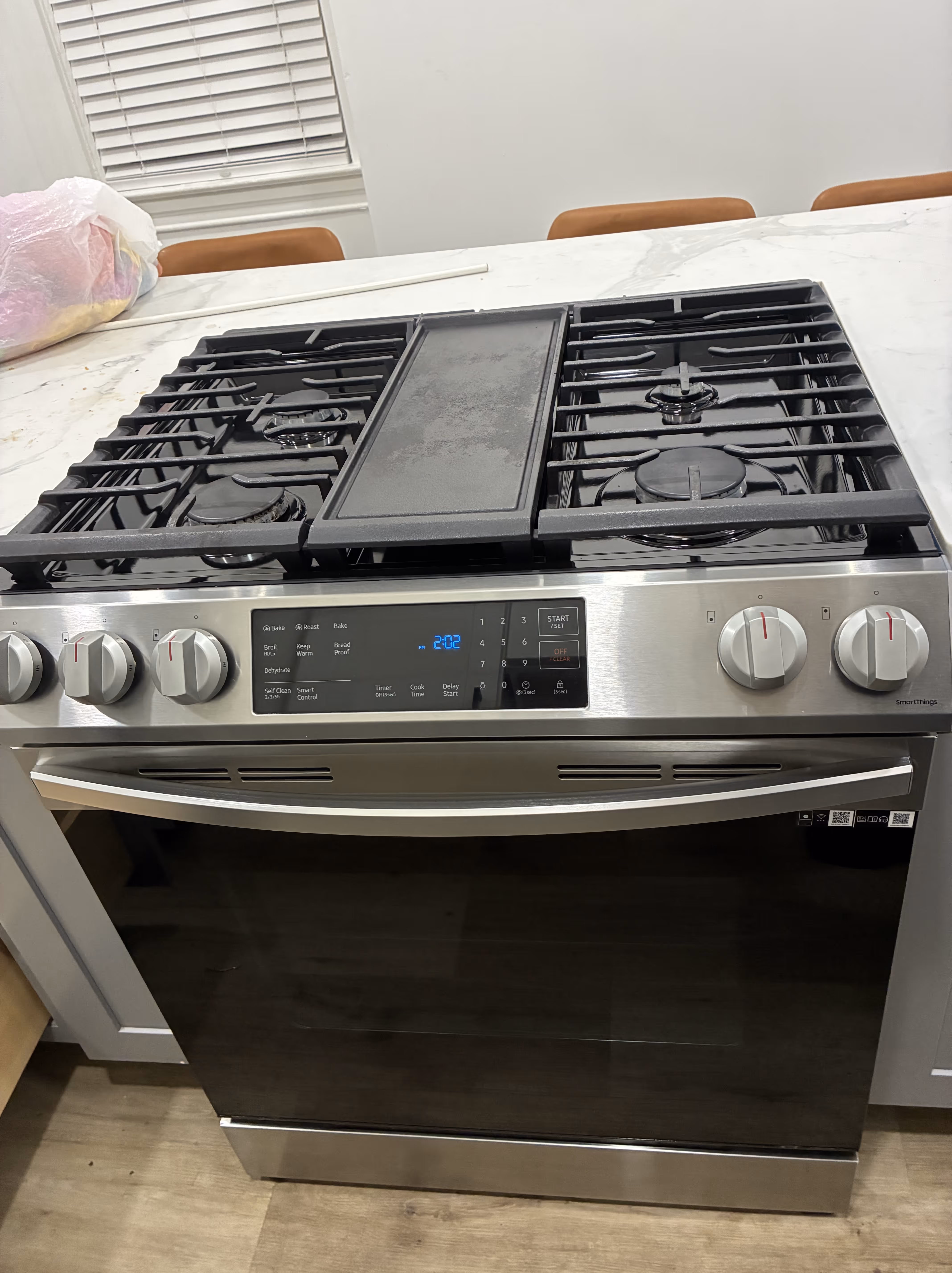 Stainless steel gas stove with black grates and digital clock displaying 9:45 with a black oven mitt hanging on the oven door handle.