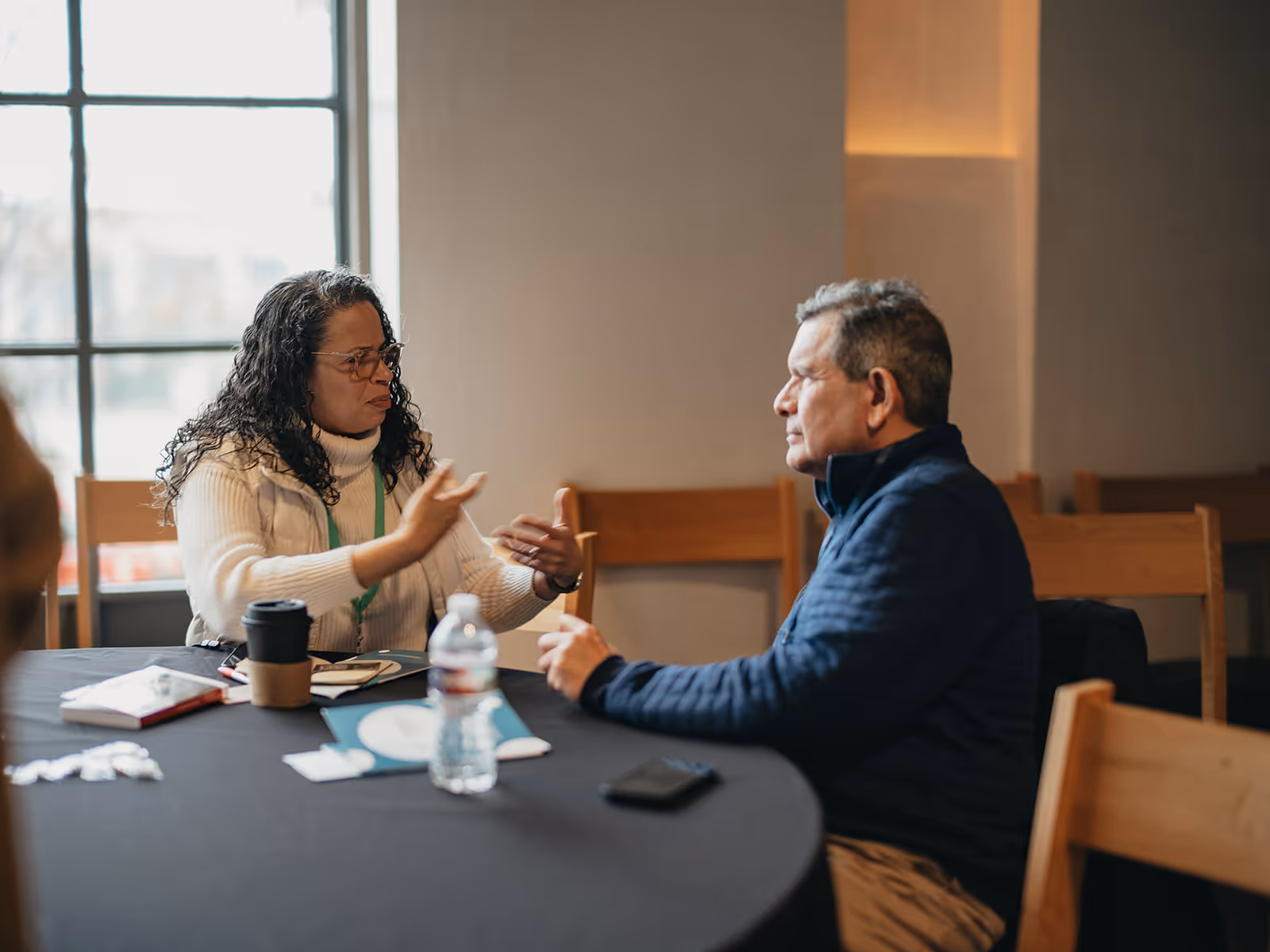 Two middle-aged individuals seated at a round table having an engaged conversation in a softly lit room.