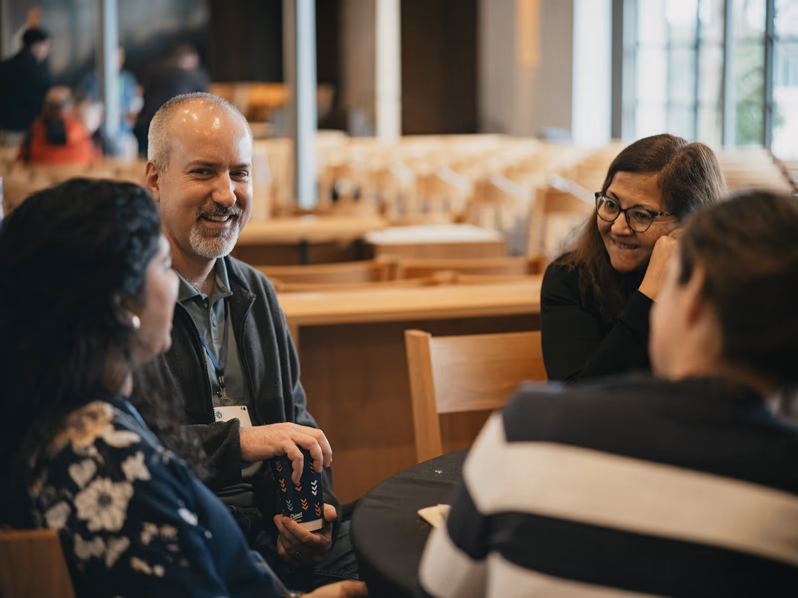 Four people having a conversation around a table in a spacious room with wooden chairs and large windows.