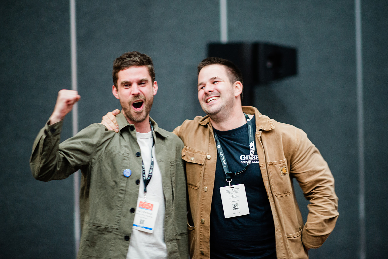 Two men smiling and posing with arms around each other at an indoor event, wearing badges on lanyards.