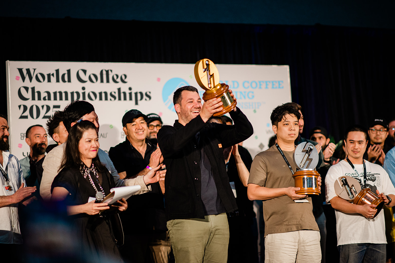 Winners at the World Coffee Championships 2025 holding trophies on stage with a group applauding behind them.