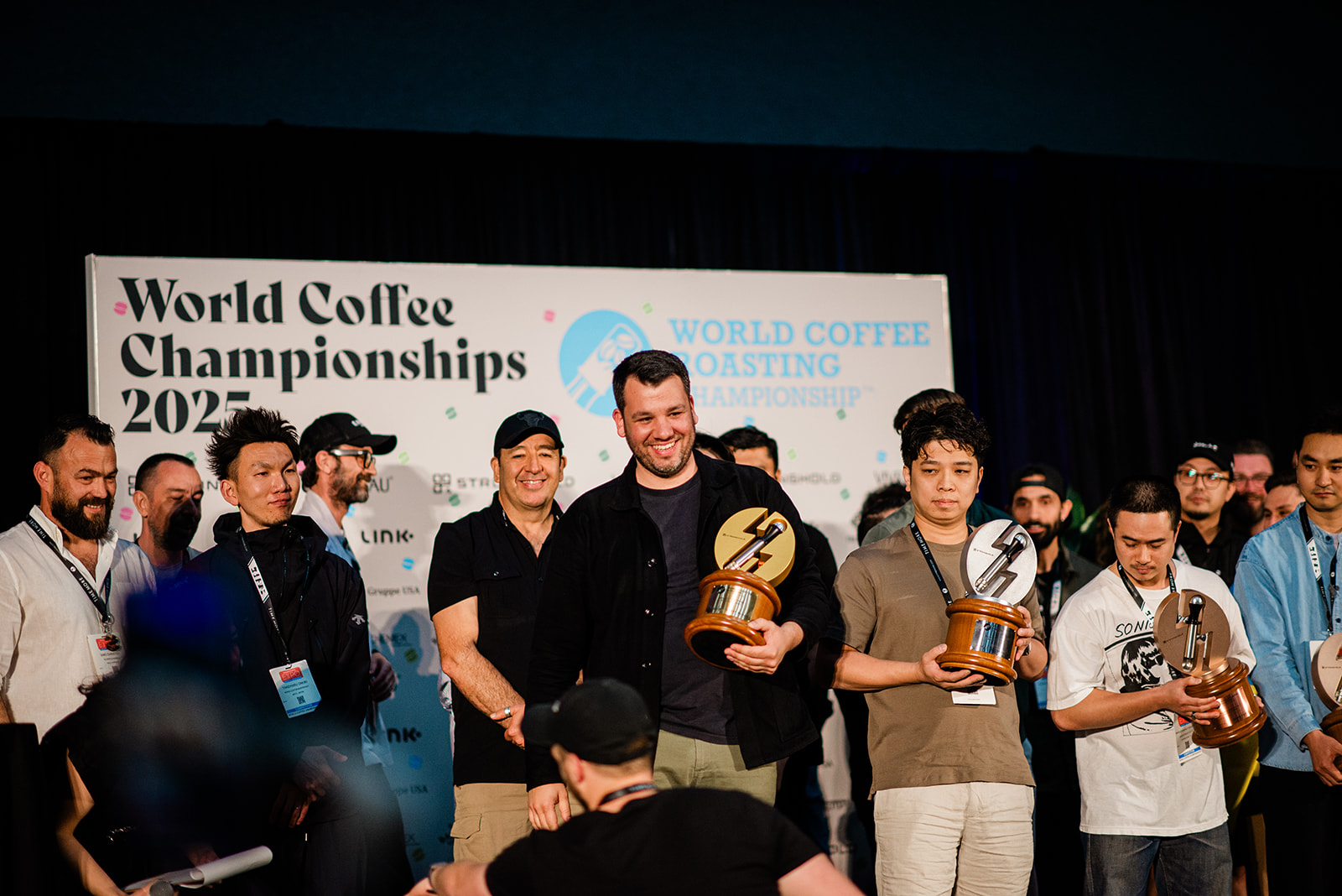 Winners holding trophies posing on stage in front of a World Coffee Championships 2025 banner.