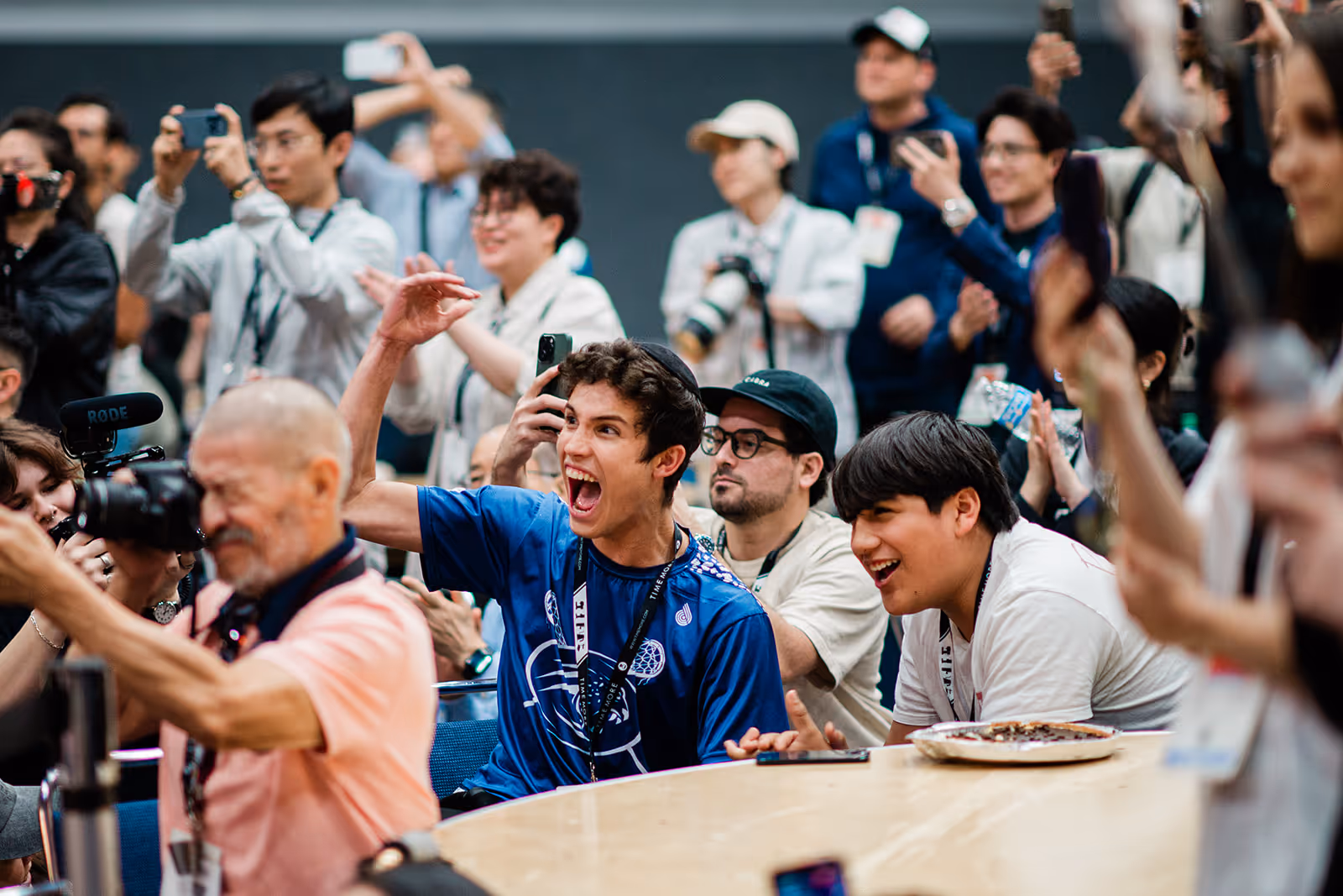 Excited young people cheering and taking photos at a lively indoor event around a table.