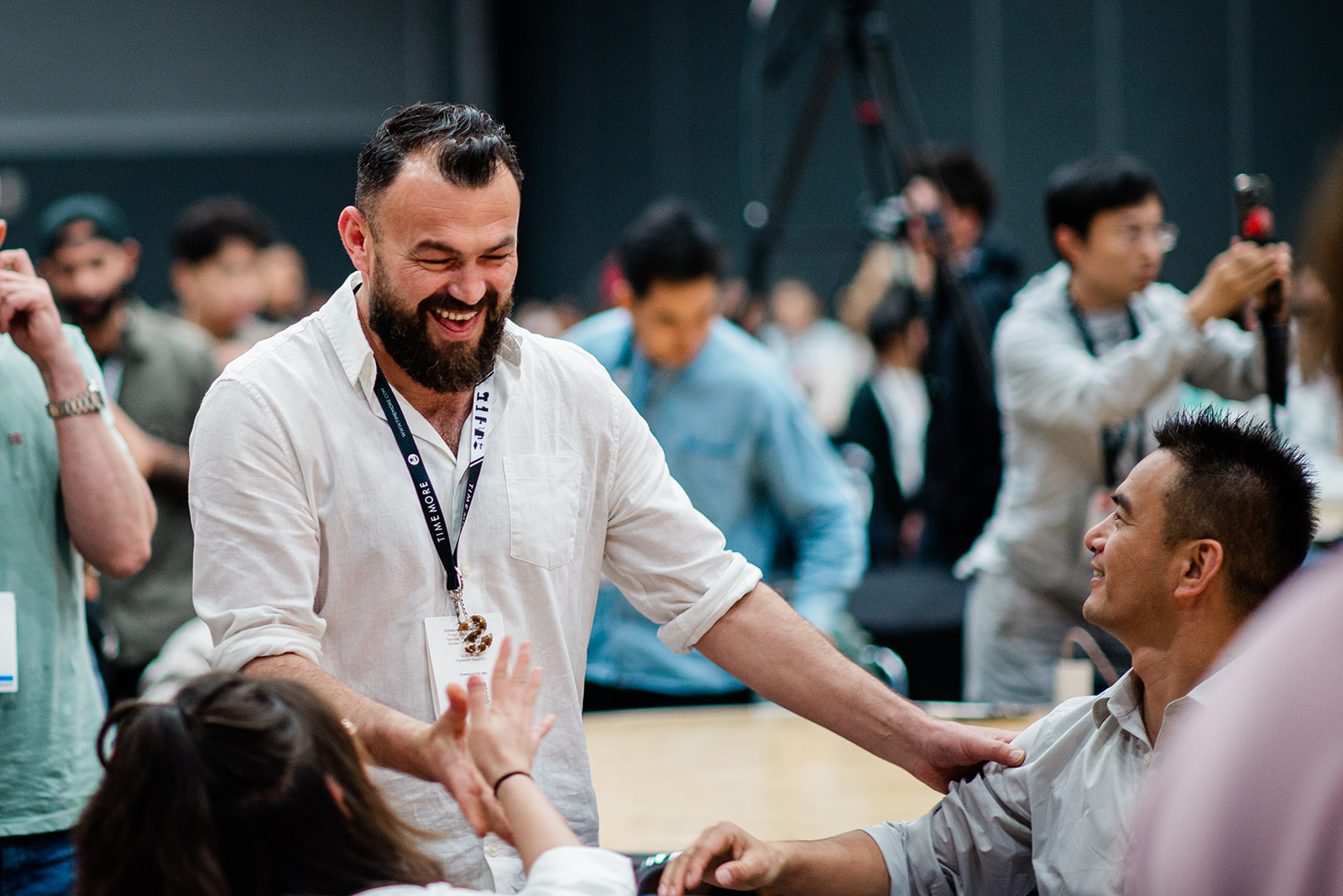 Bearded man smiling and greeting two seated people at a busy indoor event.