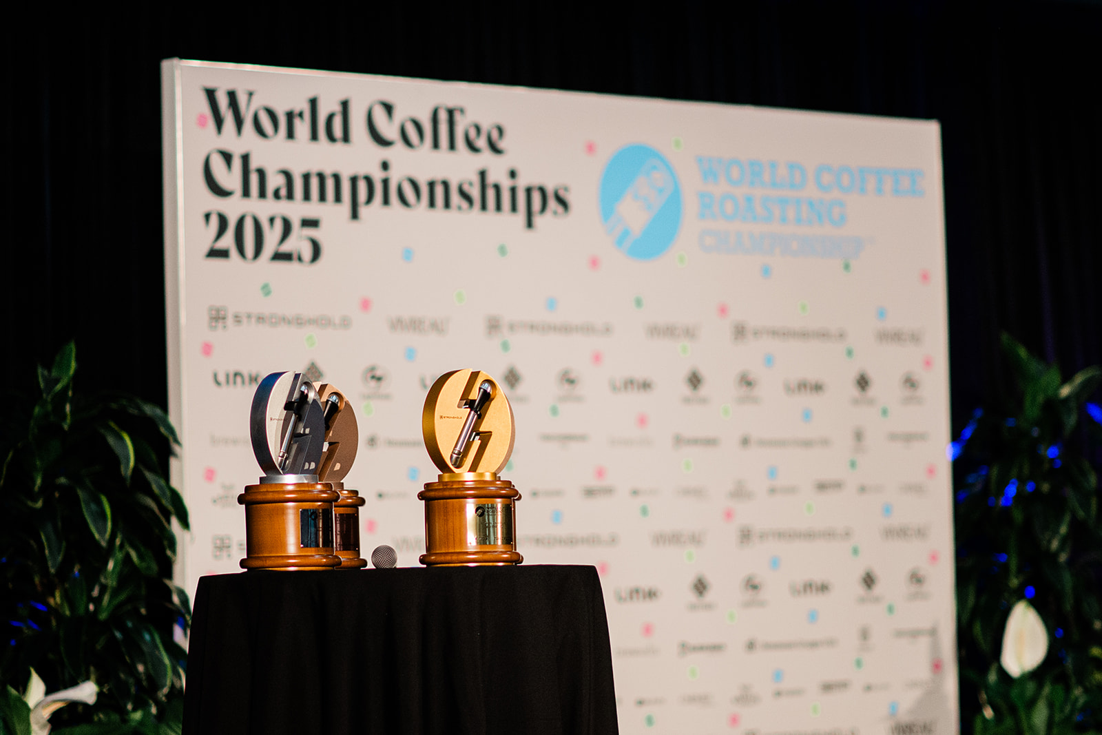 Two wooden trophies on a black-draped table in front of a World Coffee Championships 2025 banner.