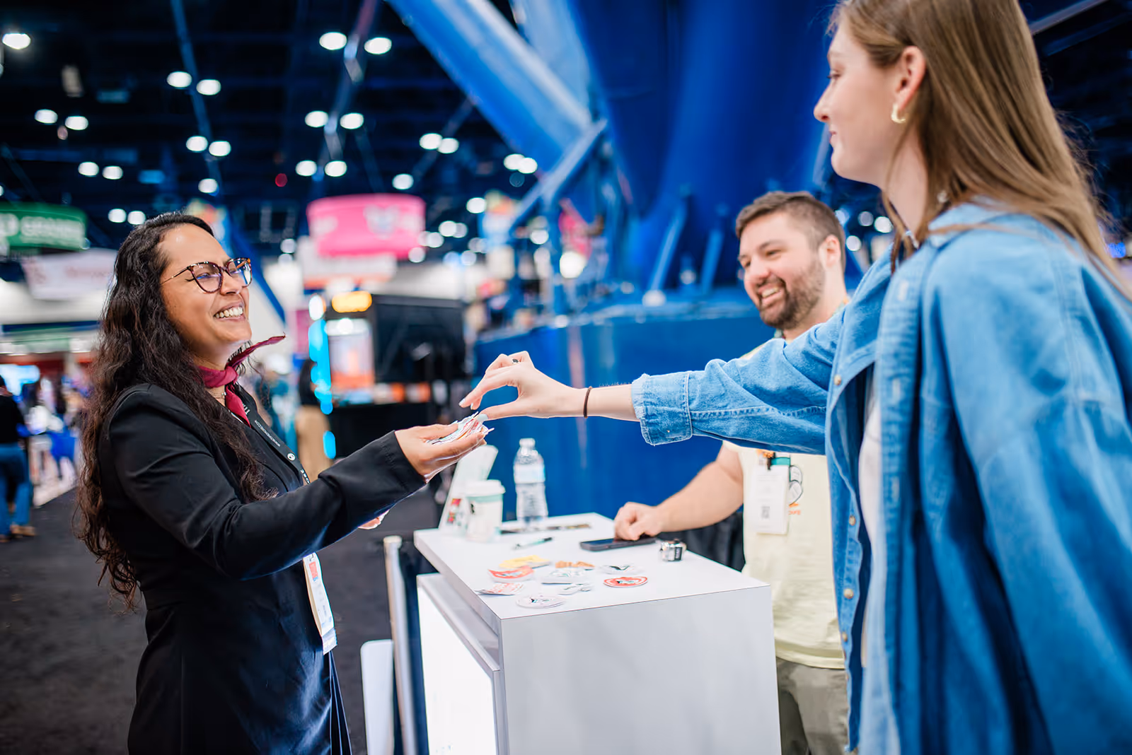 Smiling event staff woman hands stickers to a visitor in a denim jacket at a convention booth with a man smiling in the background.
