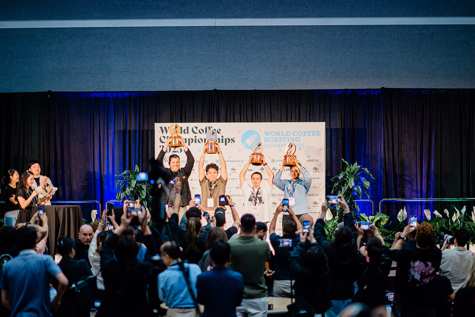 Four men on stage holding up trophies at the World Coffee Championships while an audience takes photos.