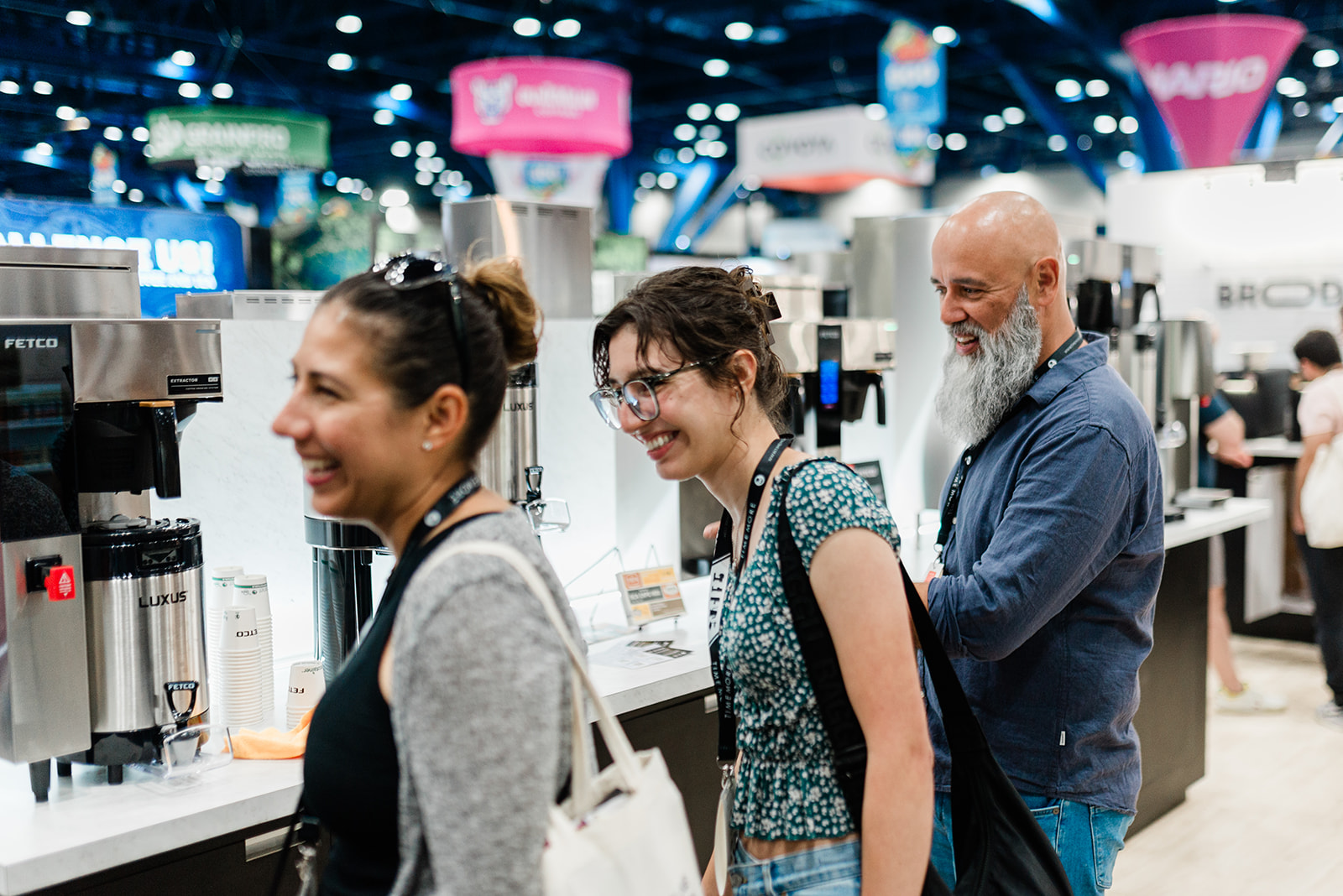 Three people smiling and standing near coffee dispensers at an indoor event or trade show.