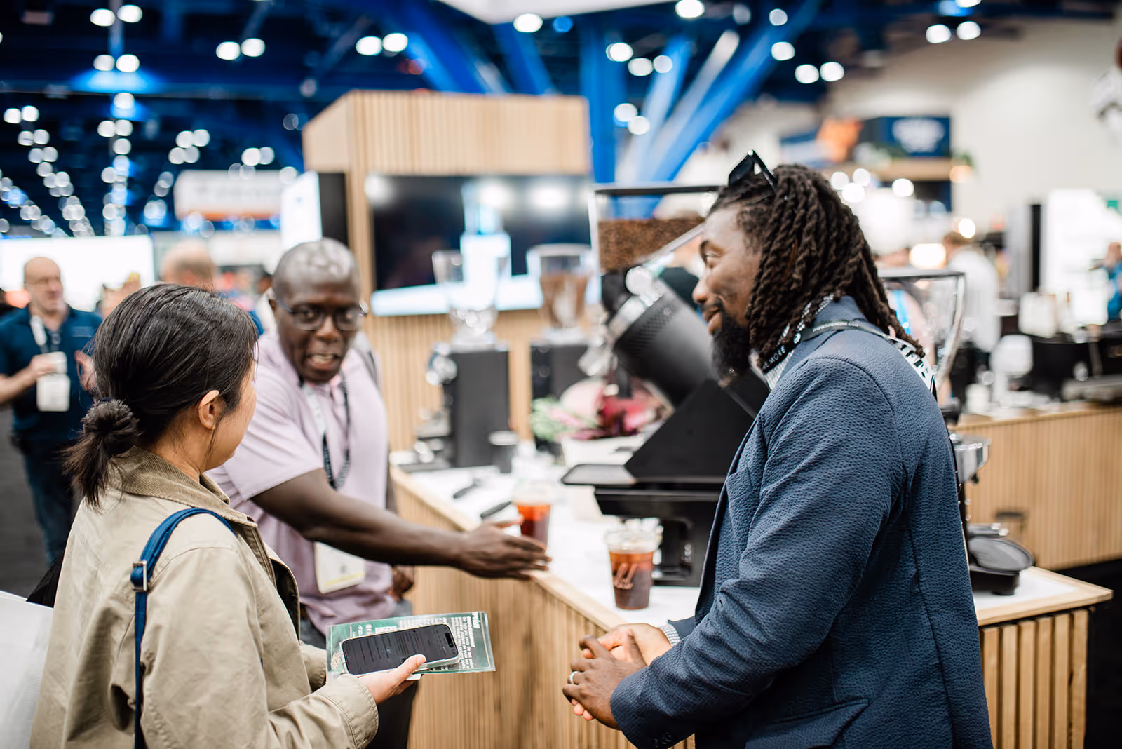 Three people engage in conversation at a coffee booth in a busy indoor event space.