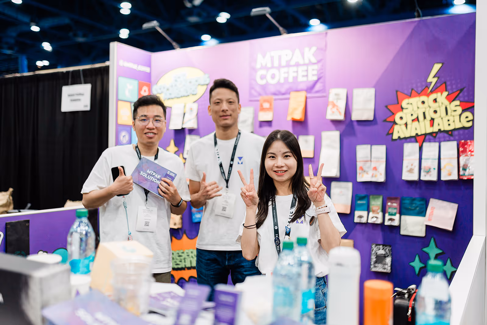 Three people wearing white shirts posing and smiling in front of a purple booth wall with various coffee packages displayed and signs reading 'MTPAK COFFEE' and 'STOCK OPTIONS AVAILABLE'.