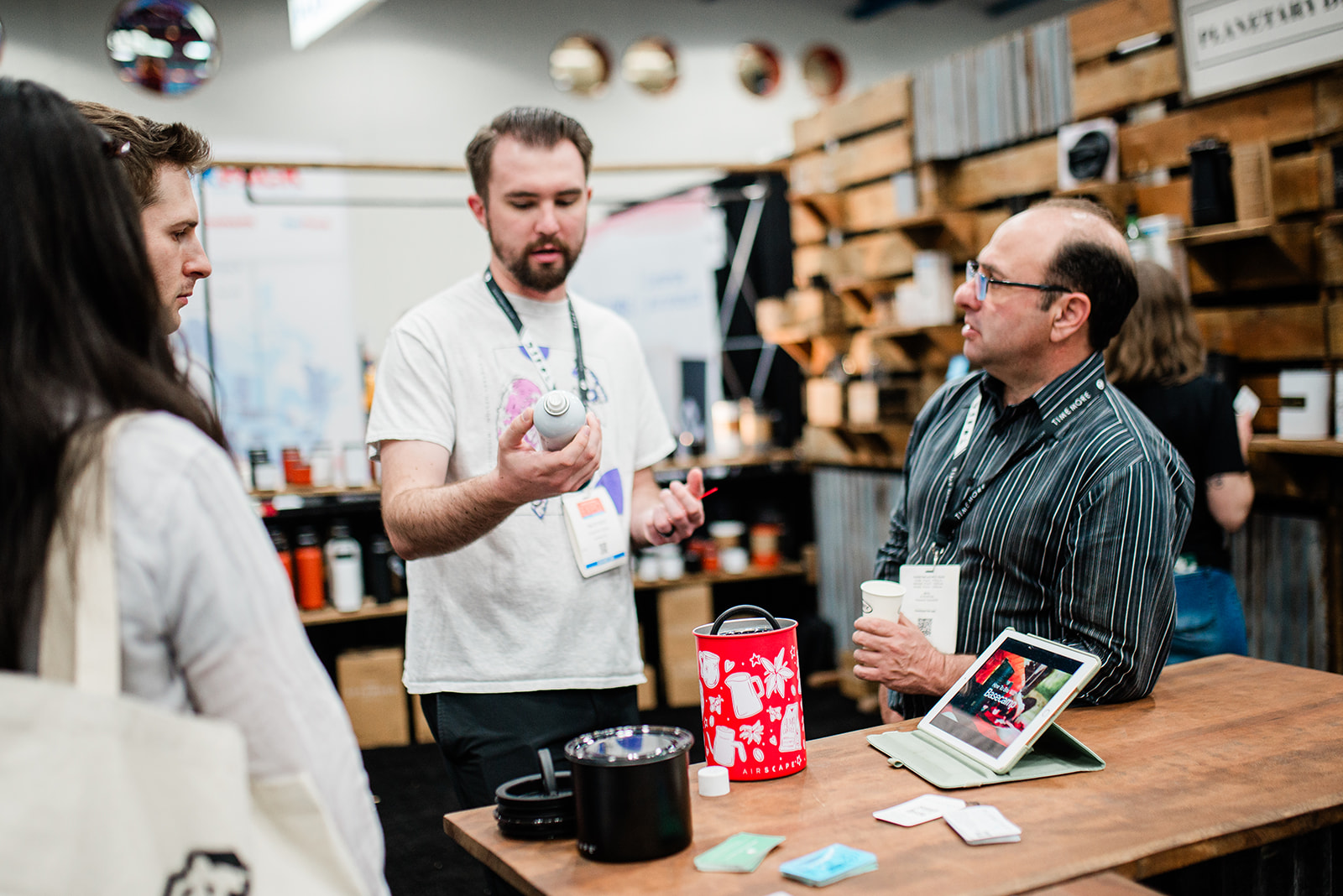 Two men and a woman engaged in conversation at a wooden display table with product samples and a tablet at an indoor exhibition booth.