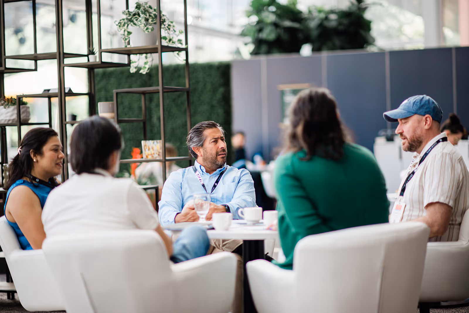 Five people engaged in a casual meeting around a small white table with coffee cups in a modern indoor space.