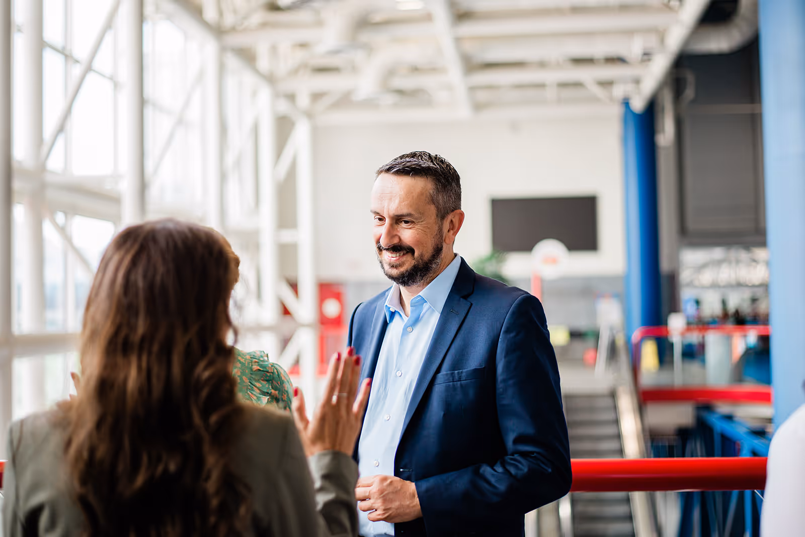 A man in a blue suit smiling and talking to a woman in a green jacket in a bright, modern indoor space.