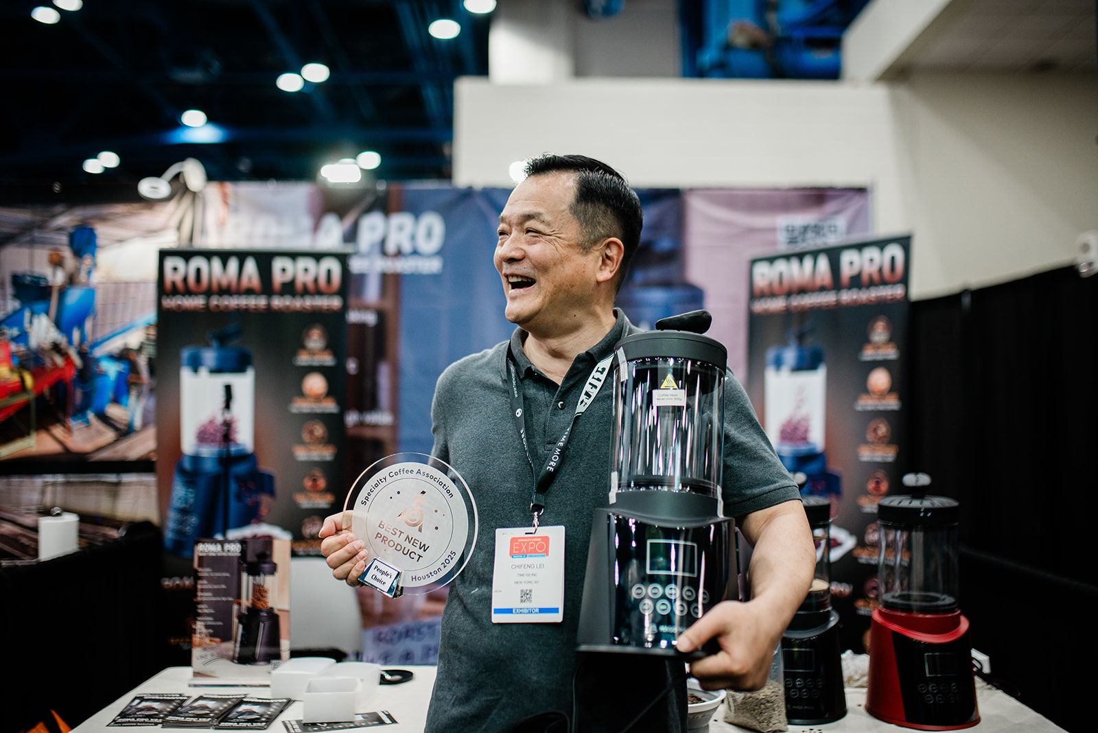 Man smiling and holding a coffee grinder and a Best New Product award plaque at a Roma Pro coffee equipment booth.